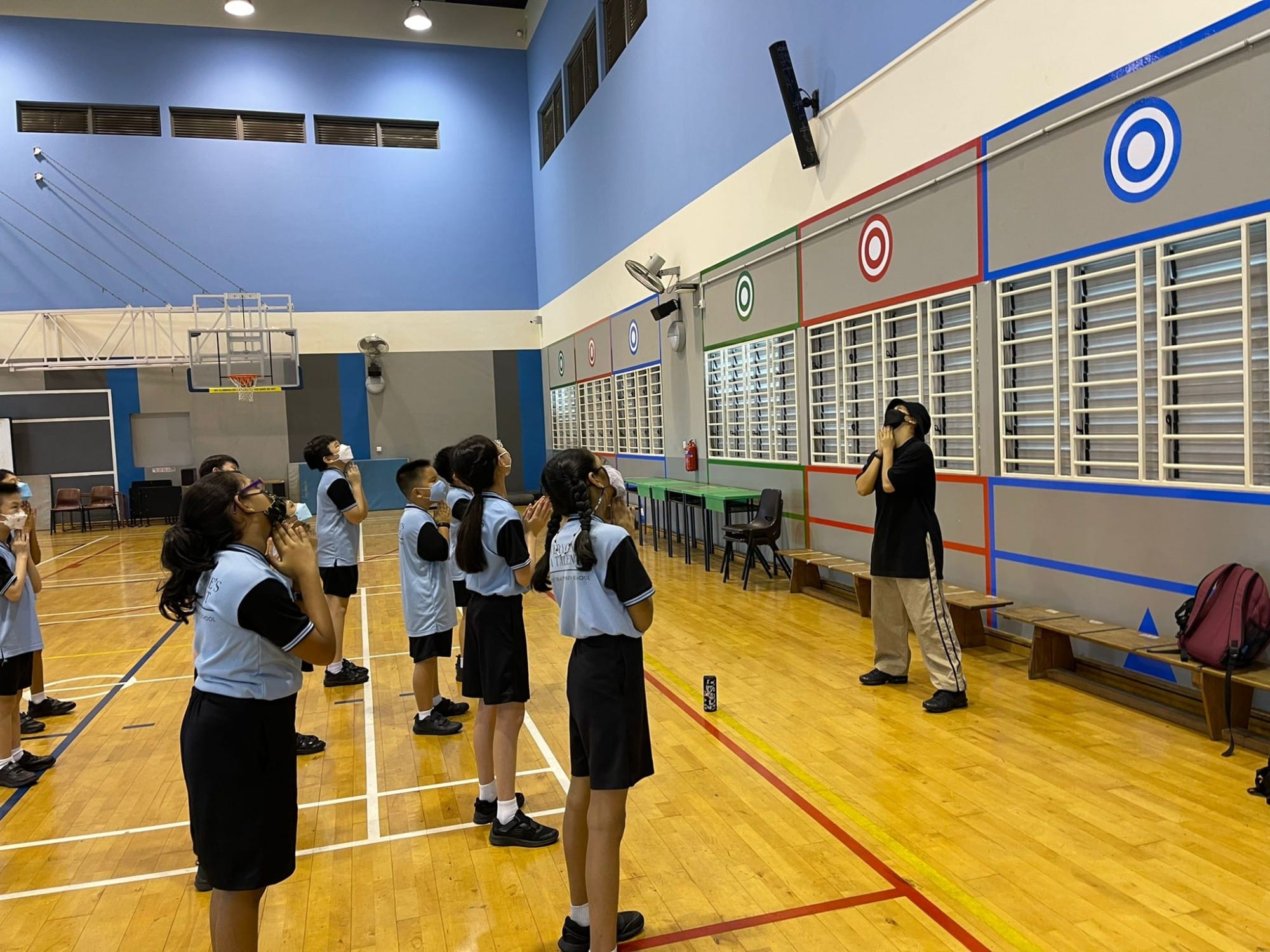 Students in light blue uniforms stand in a school gymnasium, hands raised to their faces, facing an instructor near a colorful target wall.