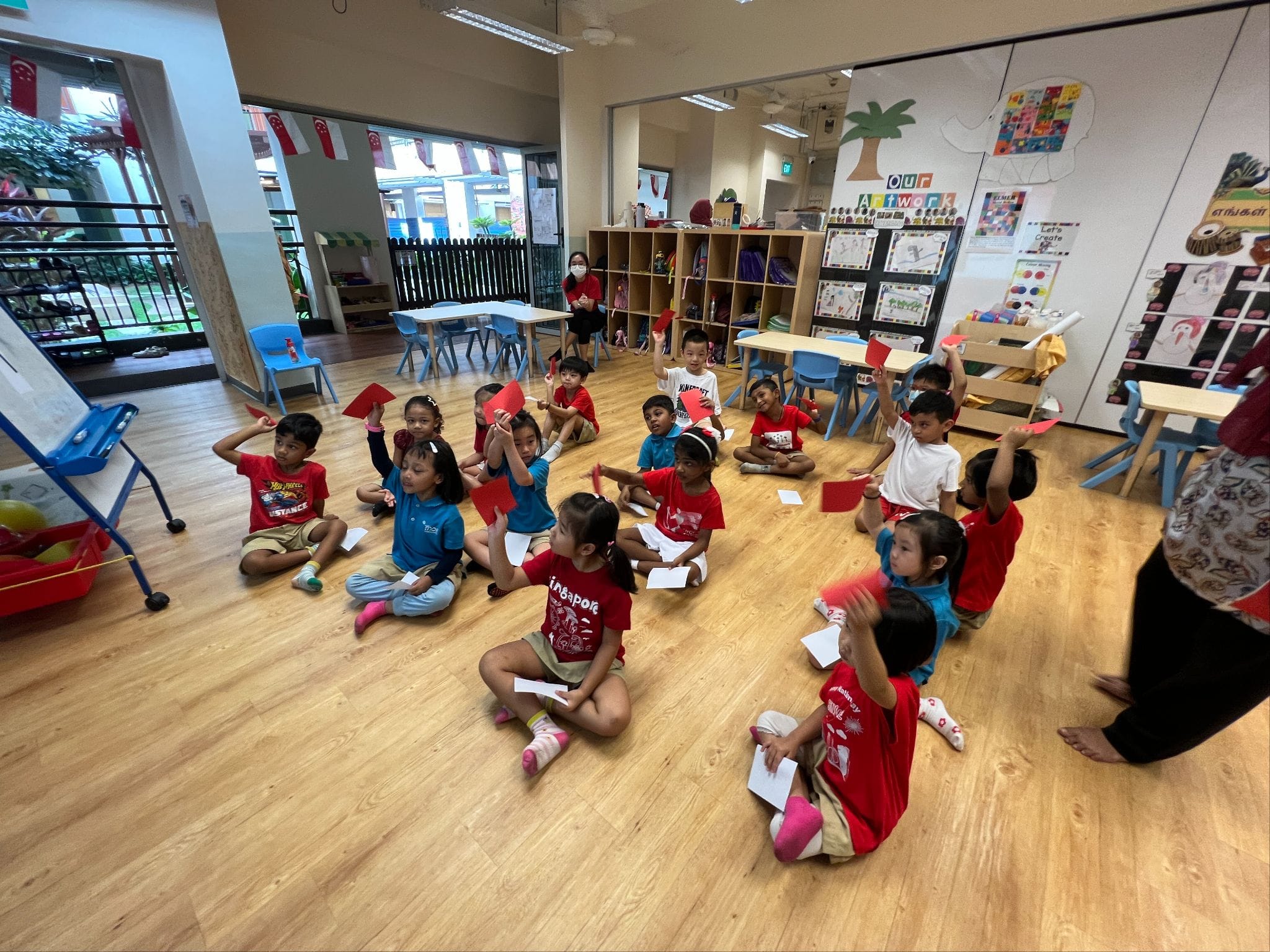 Young children sit on a classroom floor holding up red and white paper pieces, with a teacher nearby.