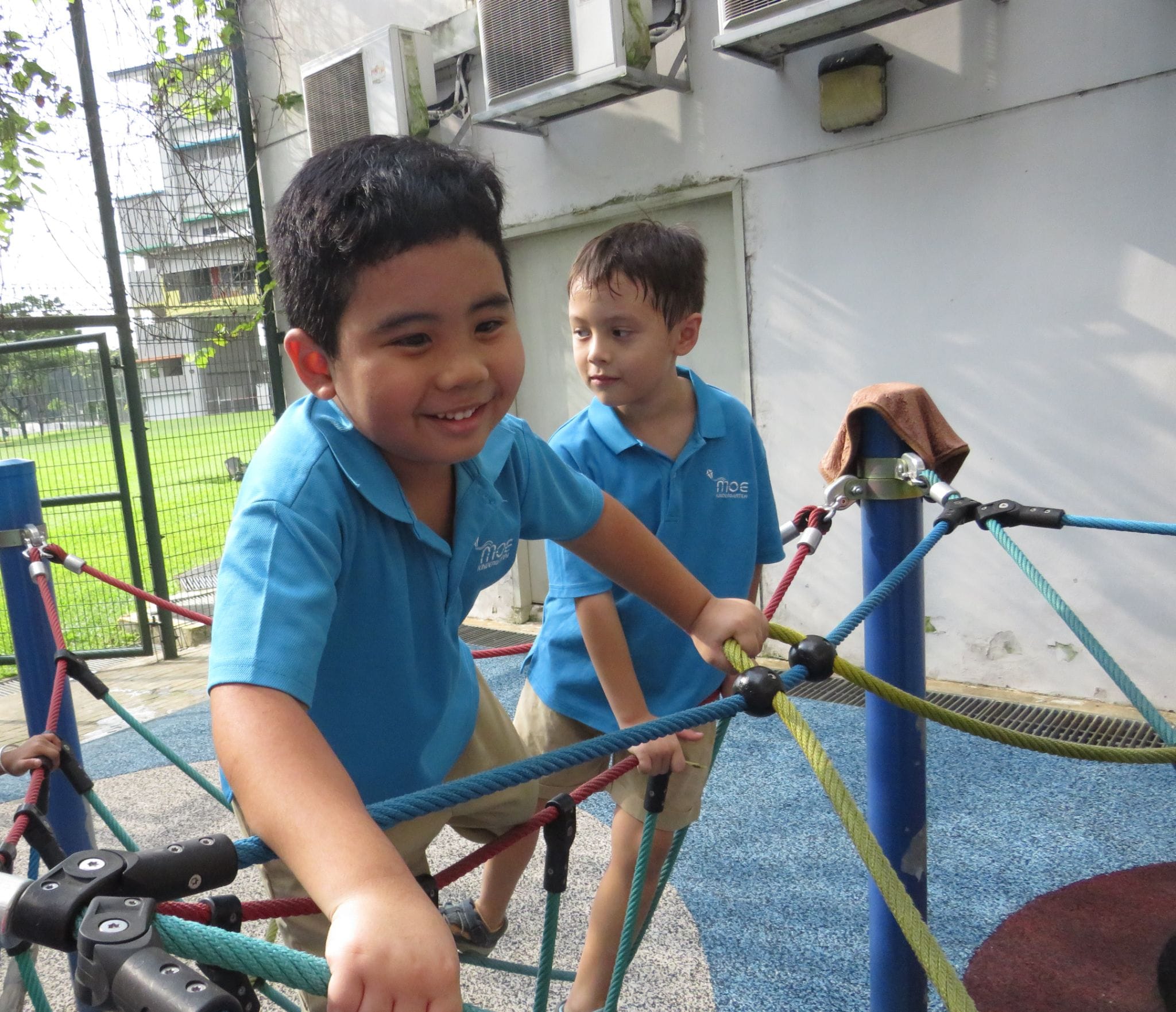 Two young boys in blue polo shirts play on a colorful rope climbing structure at an outdoor playground.