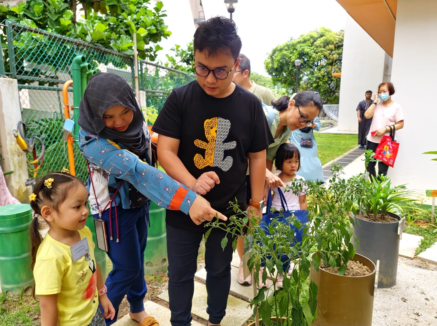 Adults and children examine potted plants outdoors, with a man pointing at green plants in large containers.