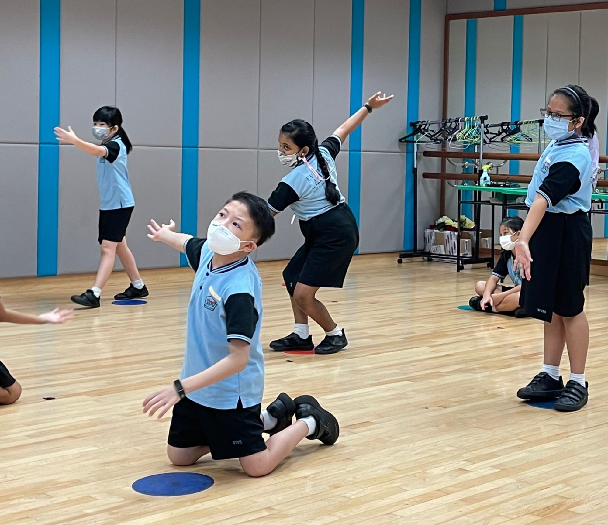 Children in blue school uniforms and face masks practice dance moves on a wooden studio floor.