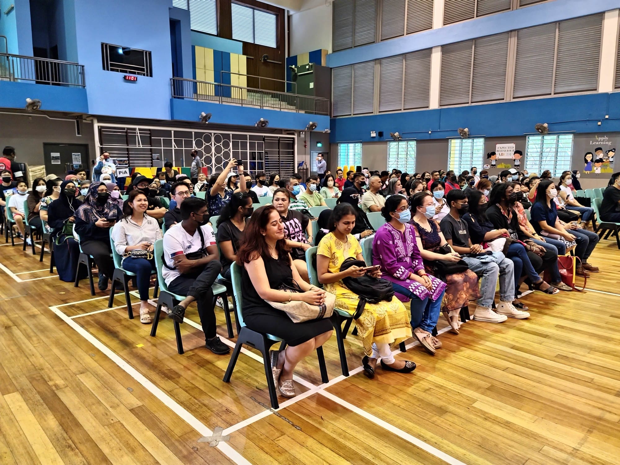A large diverse crowd sits on chairs in a school gymnasium with blue walls and a wooden floor.
