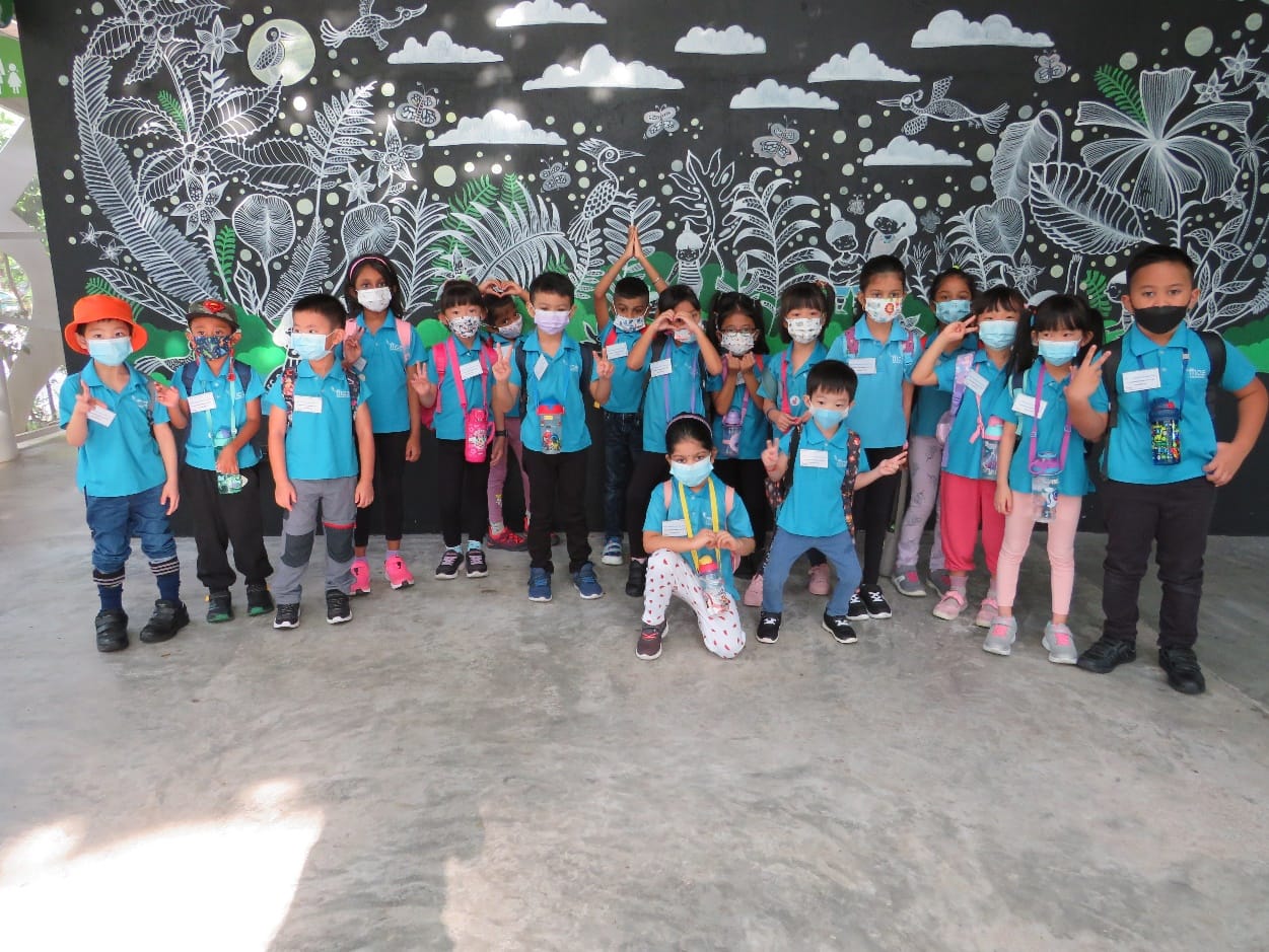A group of young children in teal uniforms and face masks pose in front of a black nature mural.