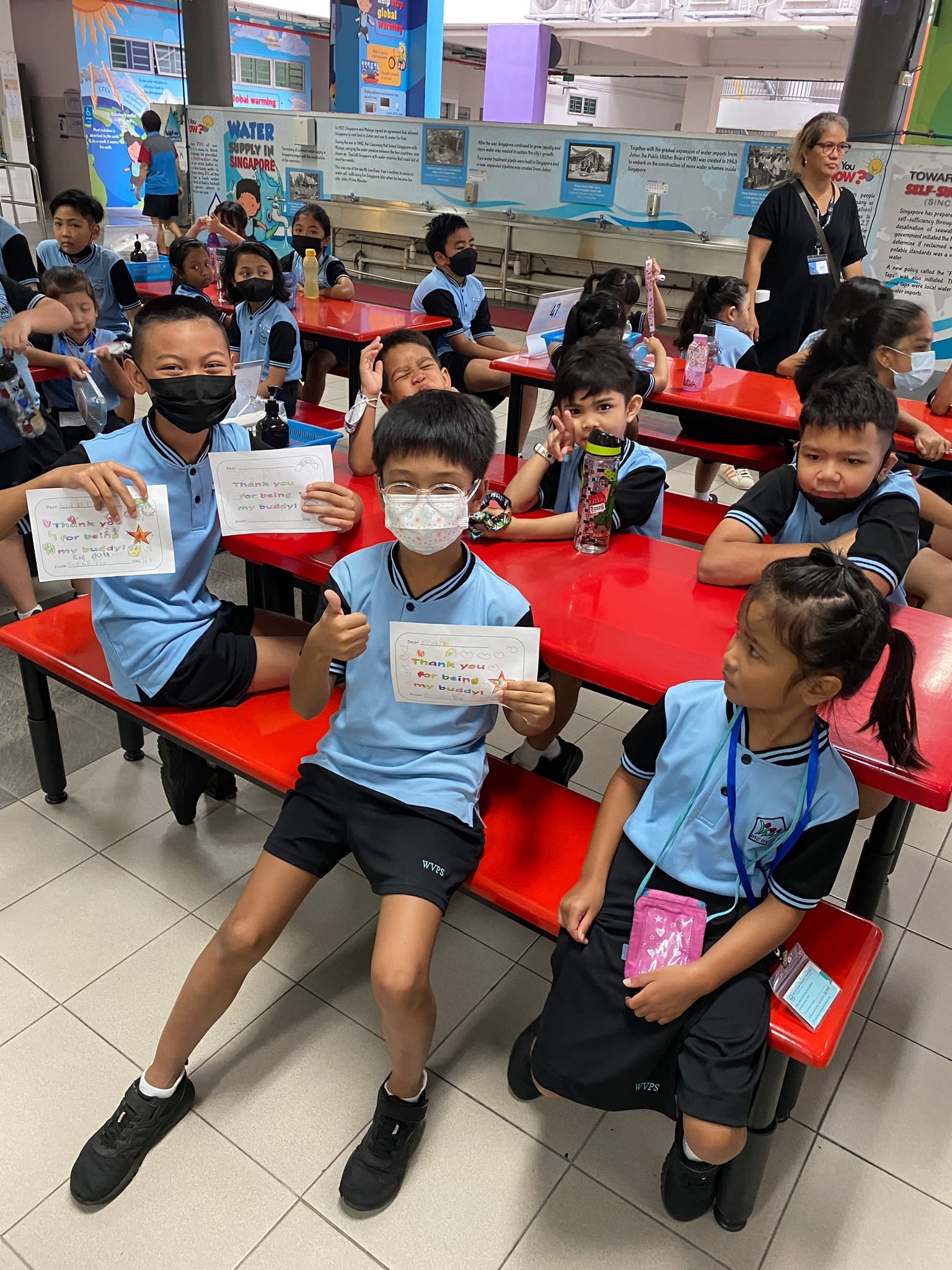 Children in blue school uniforms hold cards reading "Thank you for being my buddy" while seated at red tables.