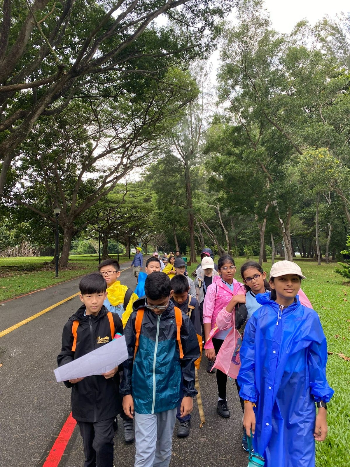 A group of children in rain jackets walk along a tree-lined park path, some carrying papers and backpacks.
