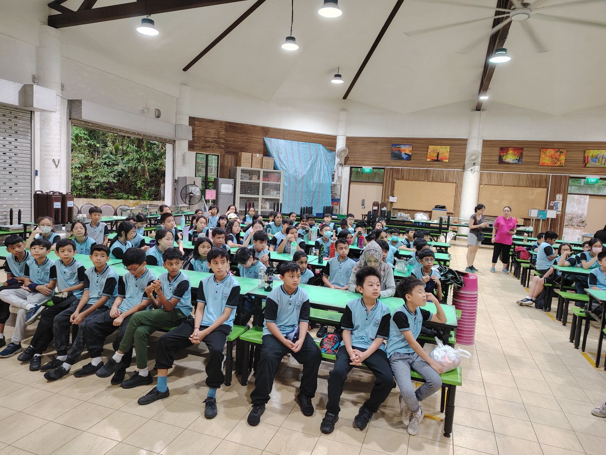 Students in blue and black uniforms sit on green benches in a school hall, facing forward attentively.