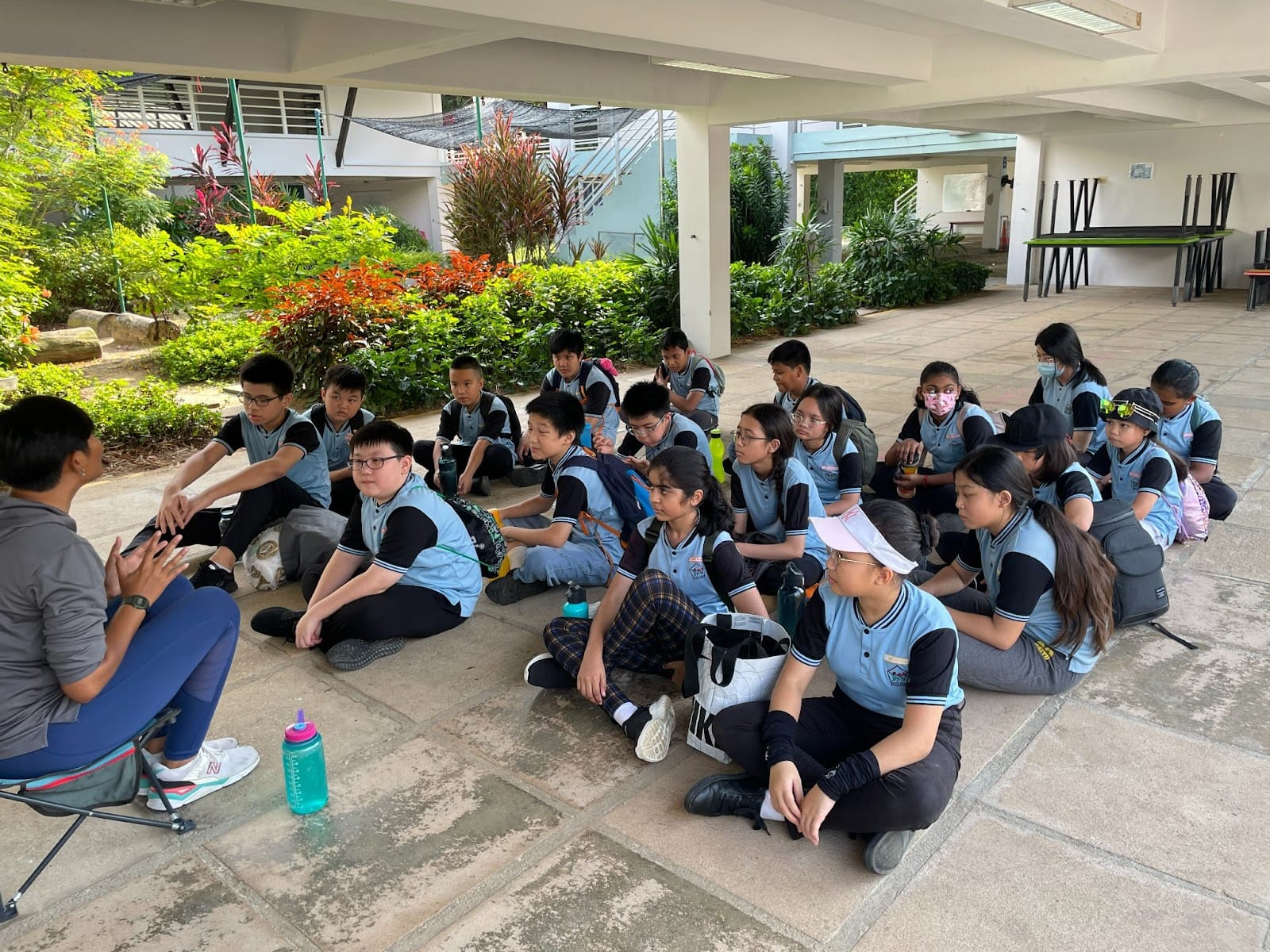 A group of students in blue uniforms sit on the ground outdoors listening to an adult instructor.
