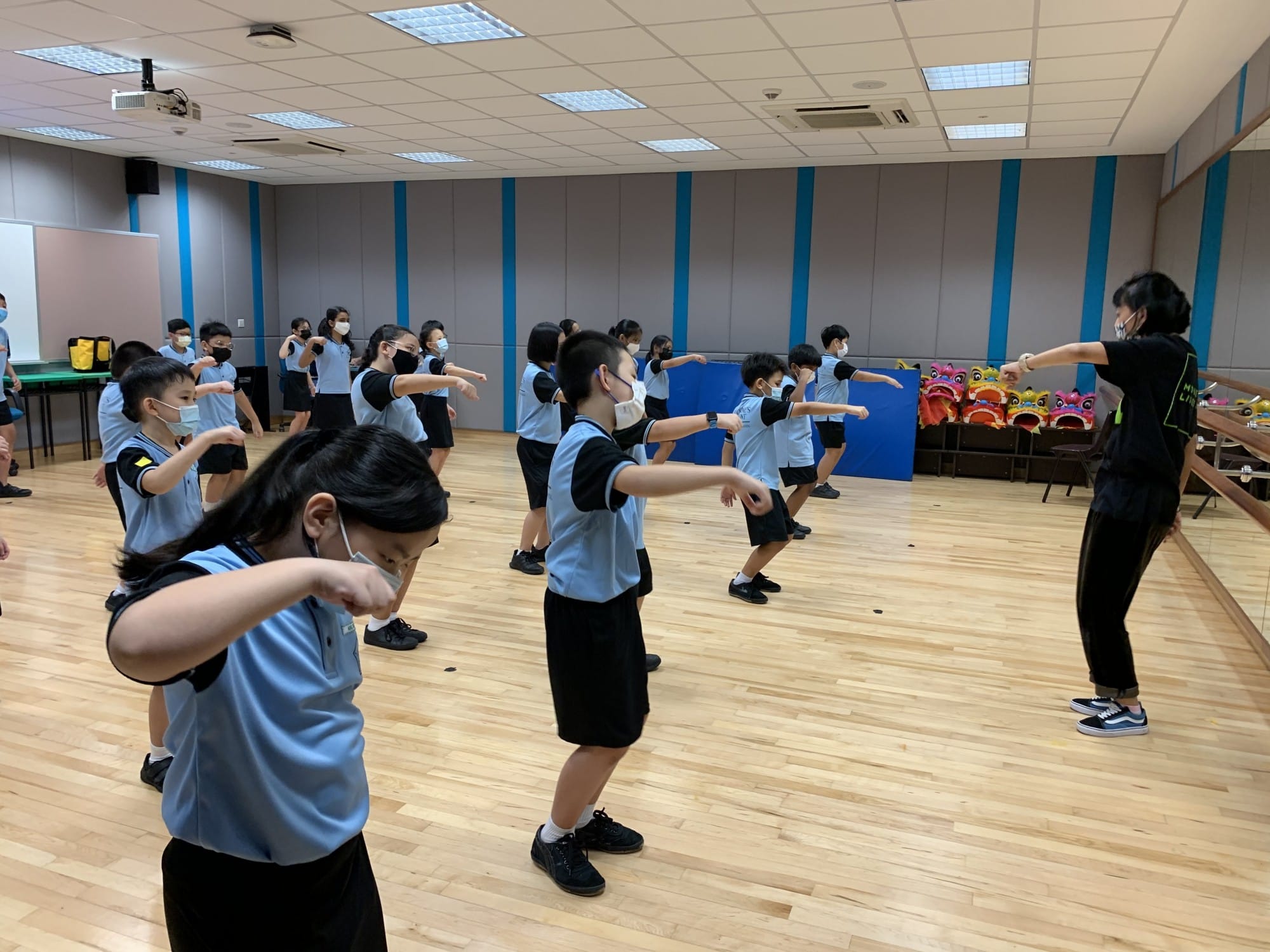 Children in blue uniforms practice martial arts moves in a studio, led by an instructor in black, with lion dance heads nearby.