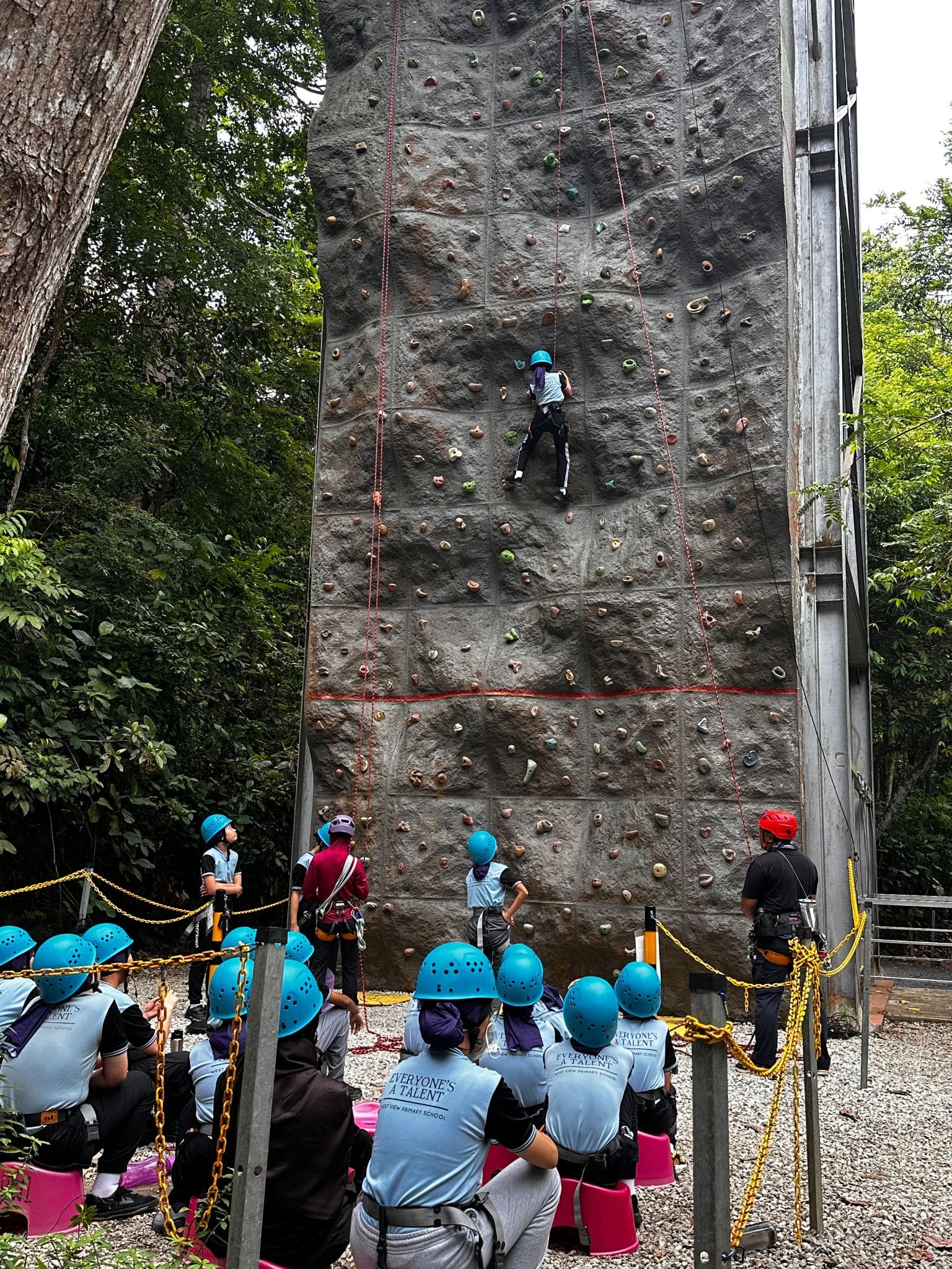 A child in a blue helmet climbs a tall outdoor rock wall while students in blue helmets watch from below.