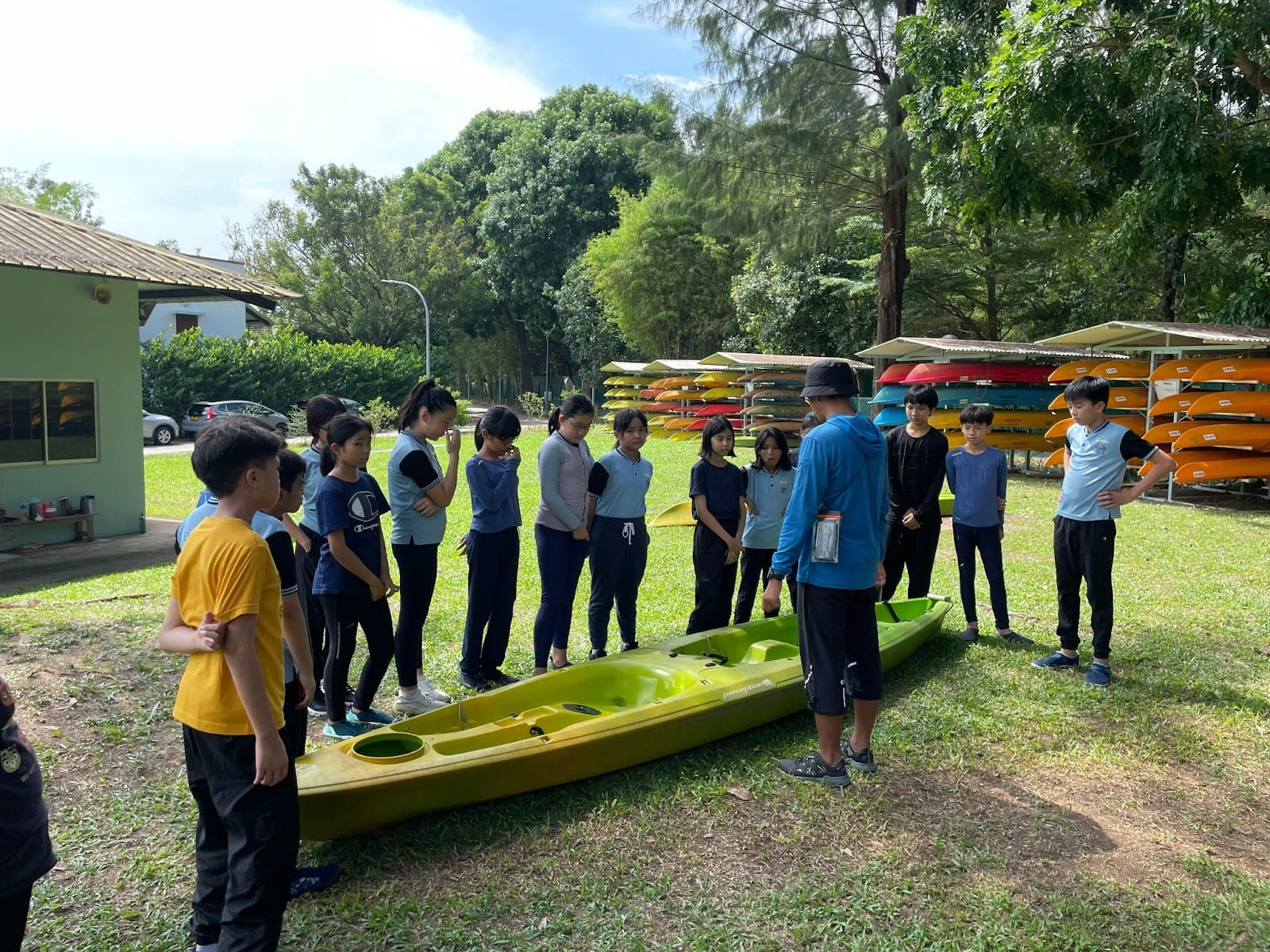 A group of children stands on grass around a green kayak while an instructor in blue speaks to them outdoors.