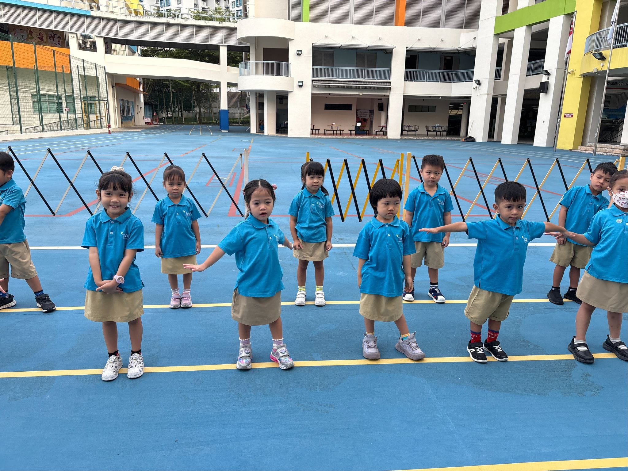 Young children in matching blue polo shirts and khaki bottoms stand in a row on a blue outdoor school court.
