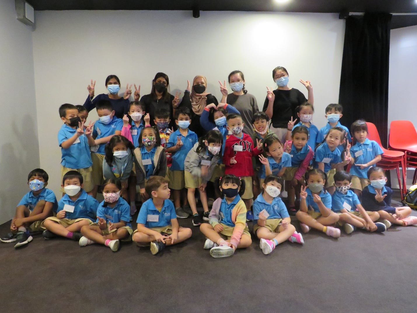 Group of young children in blue uniforms and face masks posing with adult teachers, most making peace signs.
