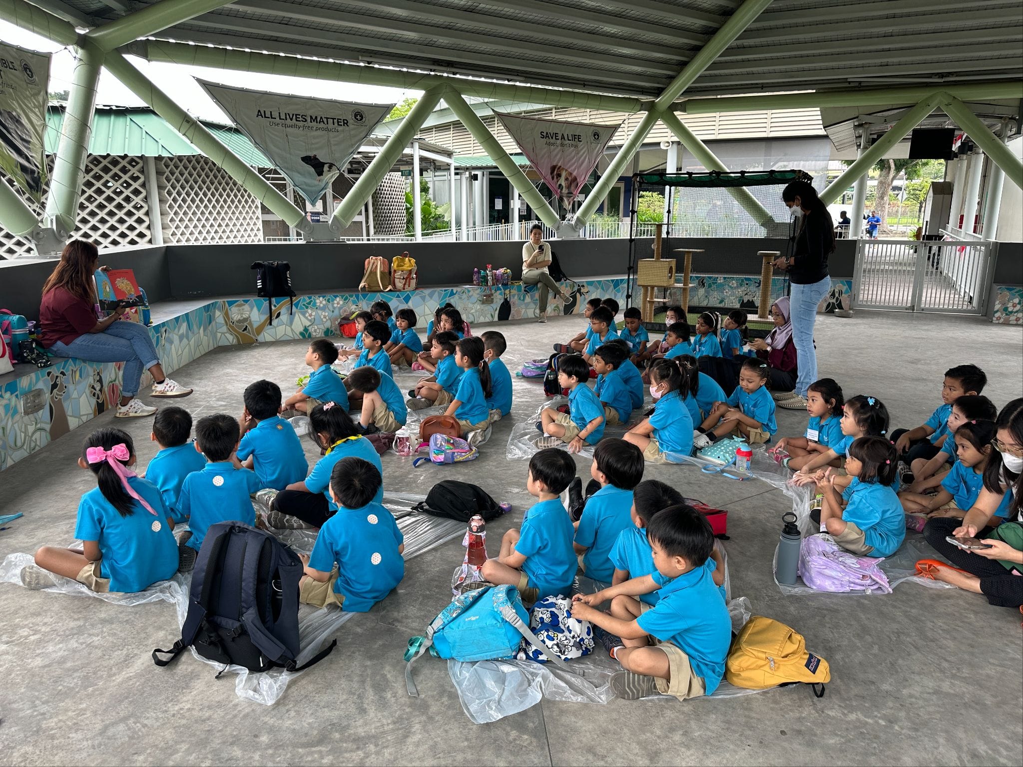 Young children in blue uniforms sit on the floor in a covered outdoor area, facing a standing adult presenter.