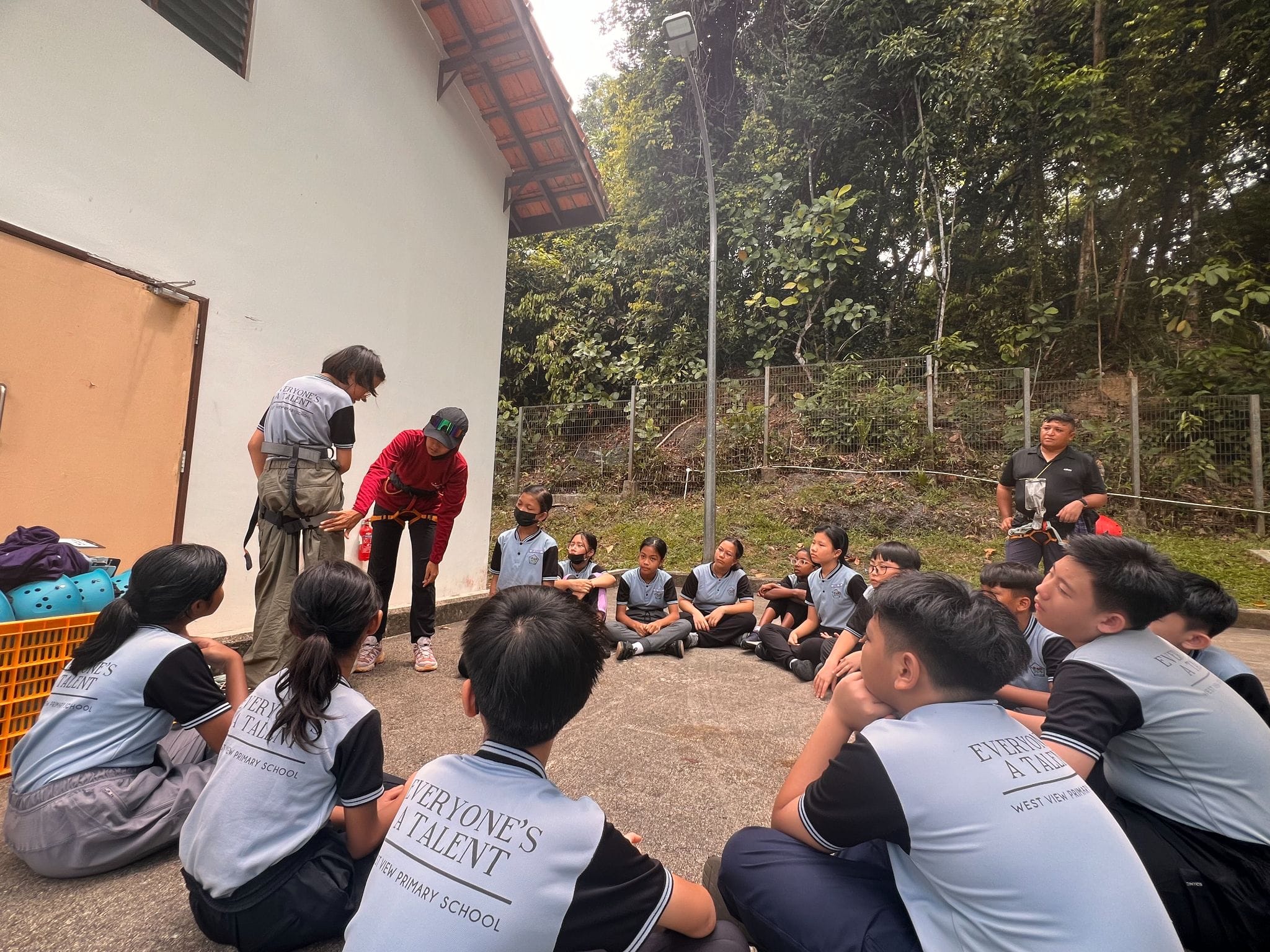Students in West View Primary School uniforms sit in a circle outdoors while two instructors address them near a building.
