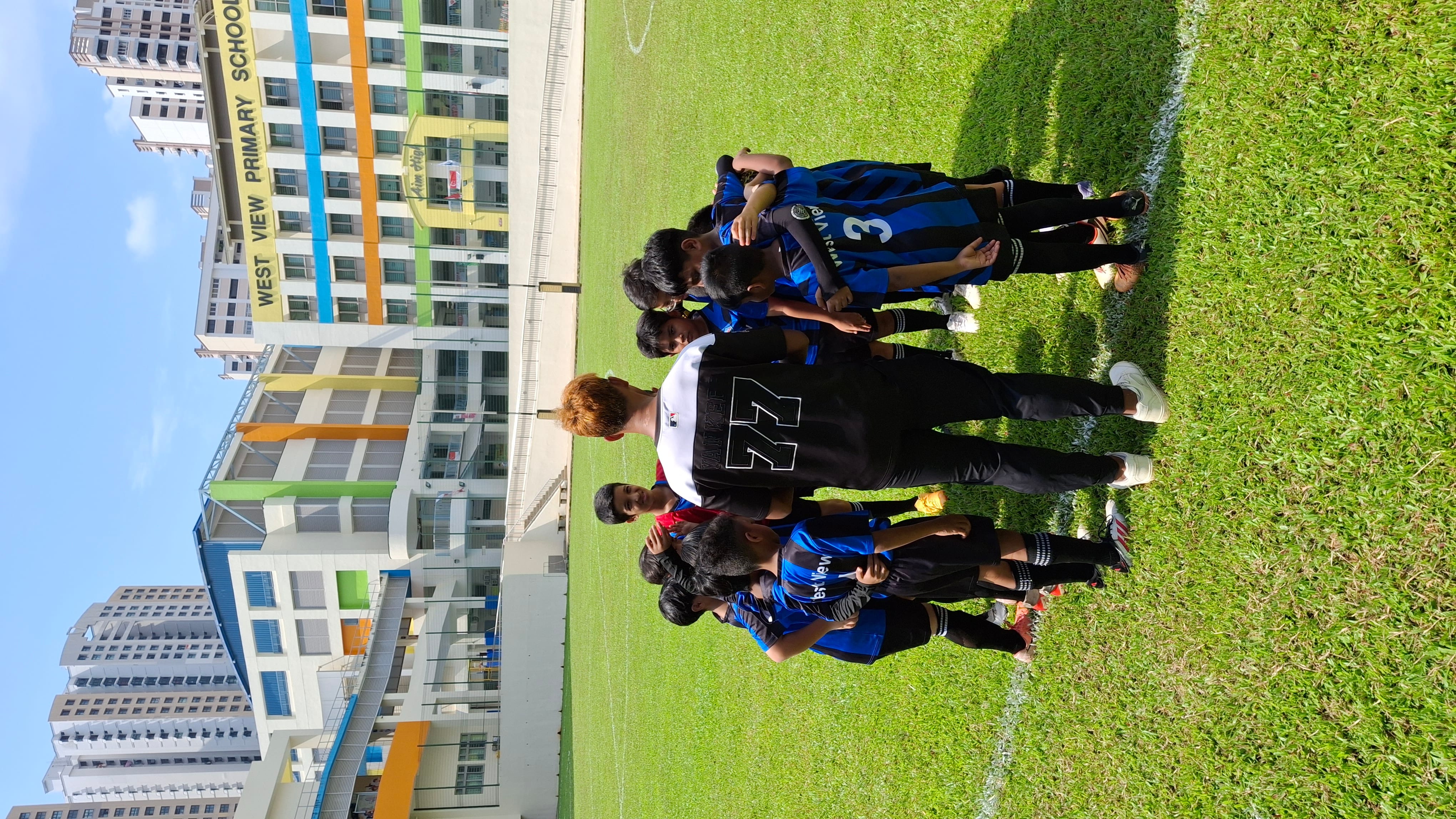 Youth soccer team huddles on grass field near a building labeled West View Primary School.