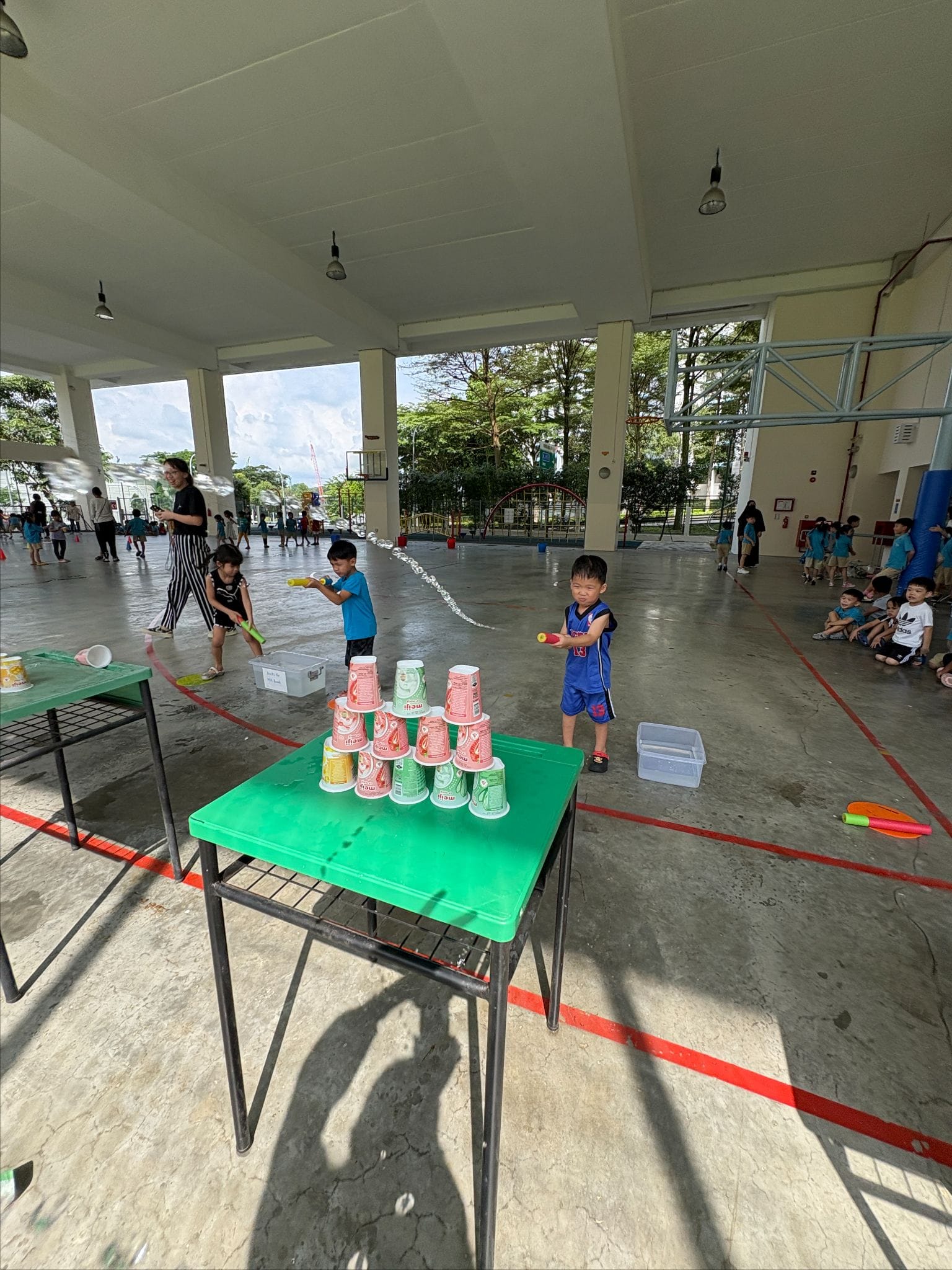 Children use water guns to knock down a pyramid of stacked cups on a green table in an open pavilion.