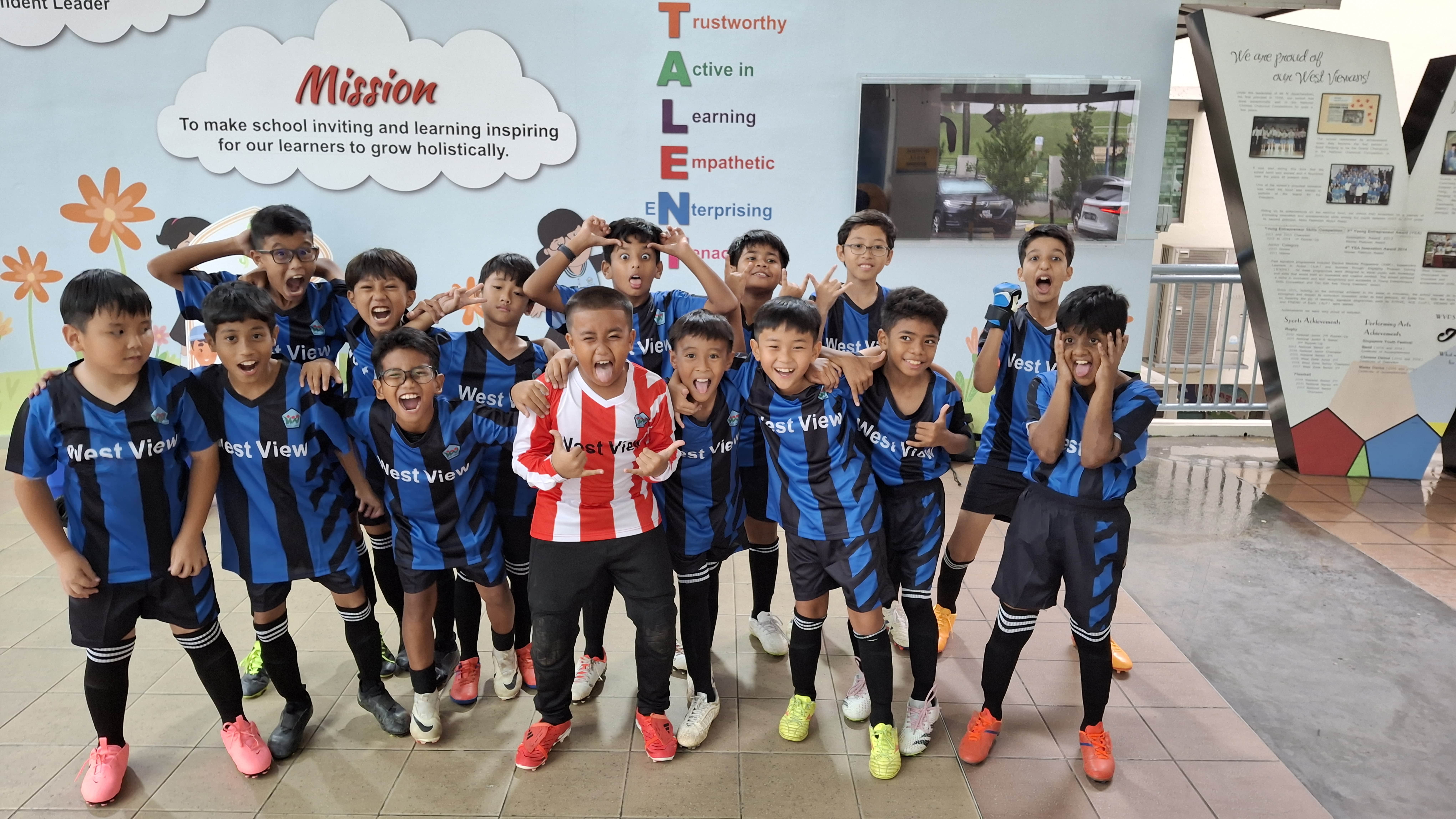 Group of boys in blue-and-black soccer kits pose excitedly indoors; one in red-and-white stands front.