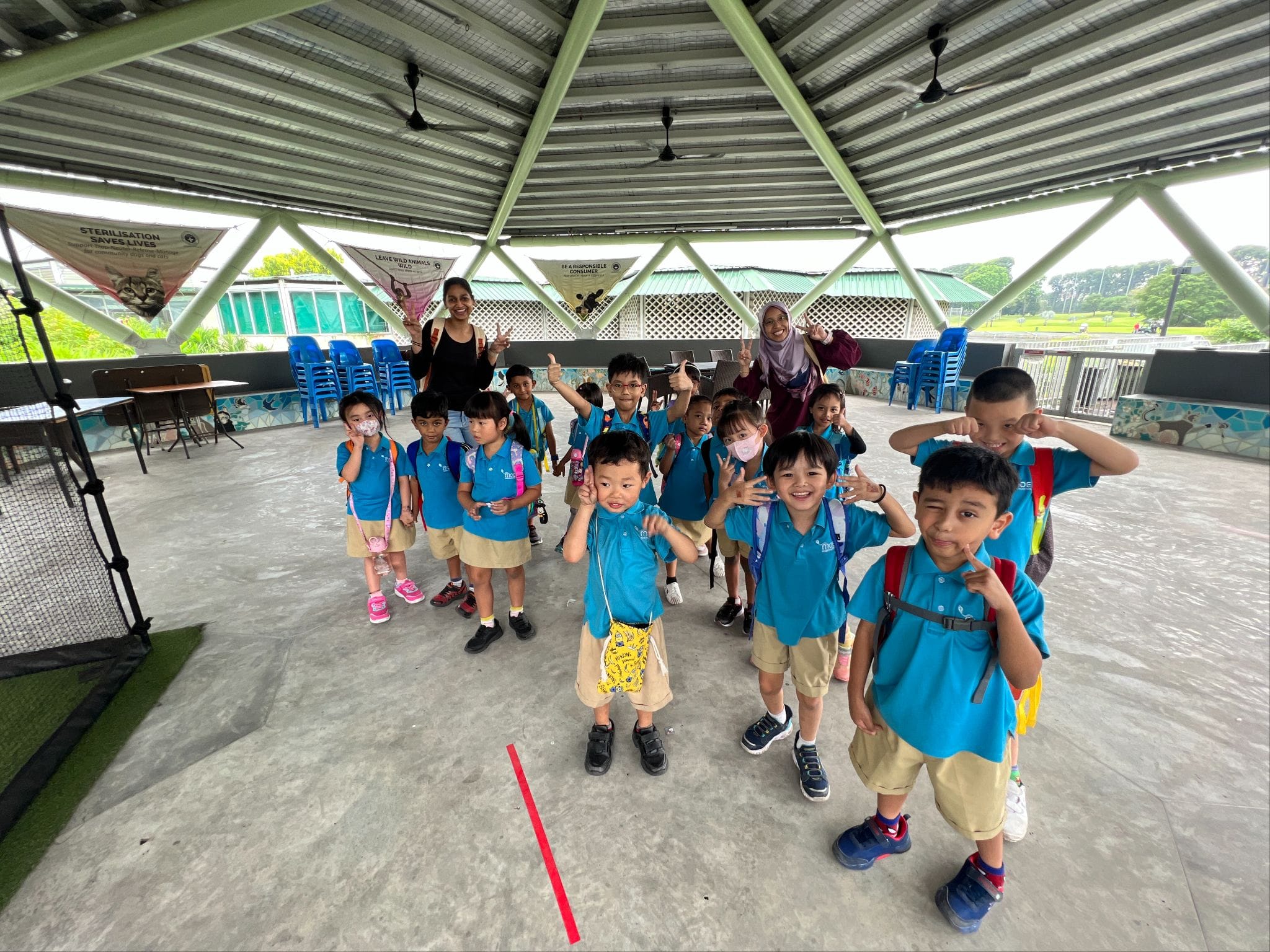 Young children in blue uniforms with backpacks pose and smile under an open pavilion with two adult chaperones.