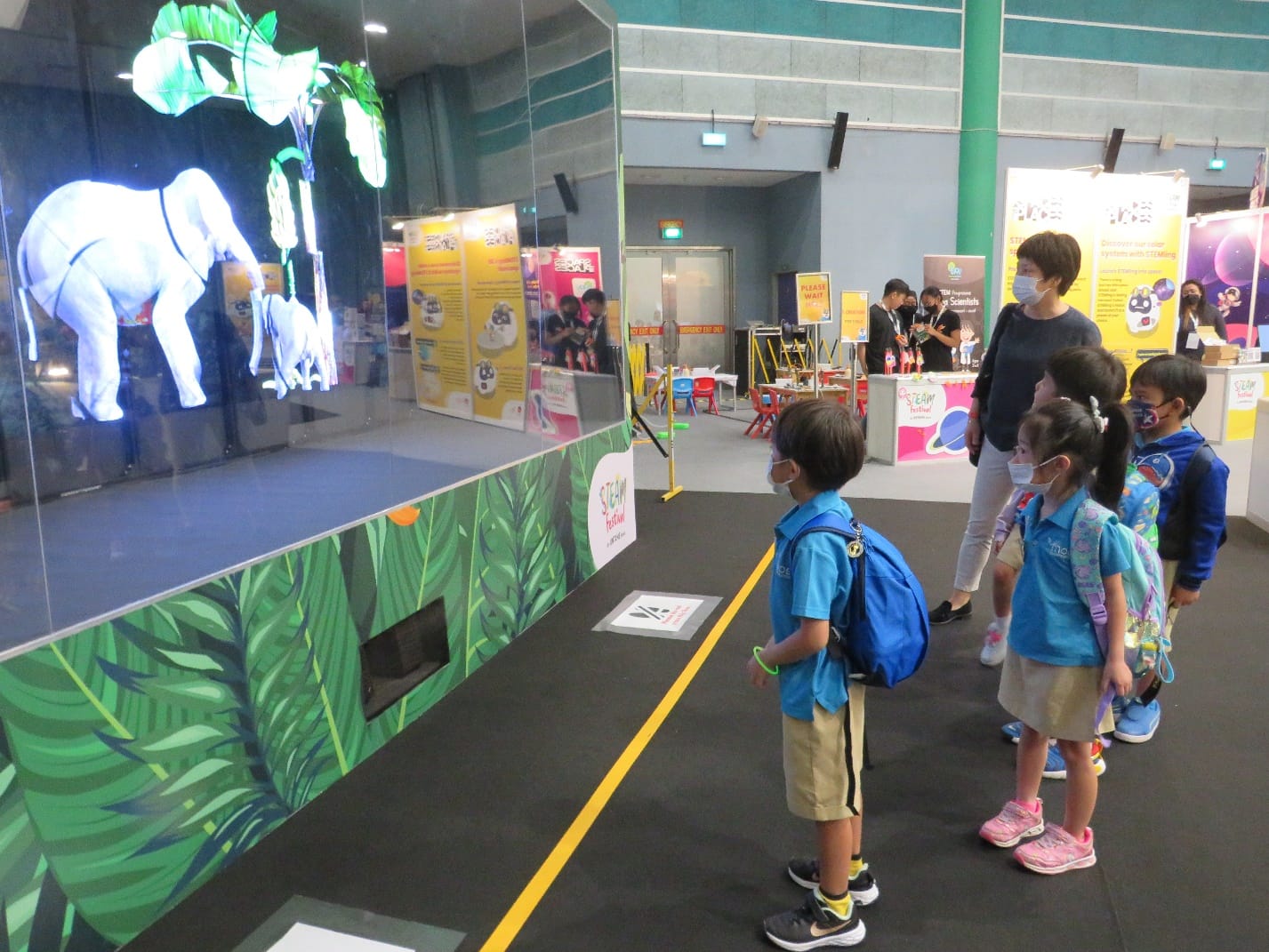 Children and an adult wearing masks watch a glowing holographic elephant display at an indoor STEAM festival exhibit.