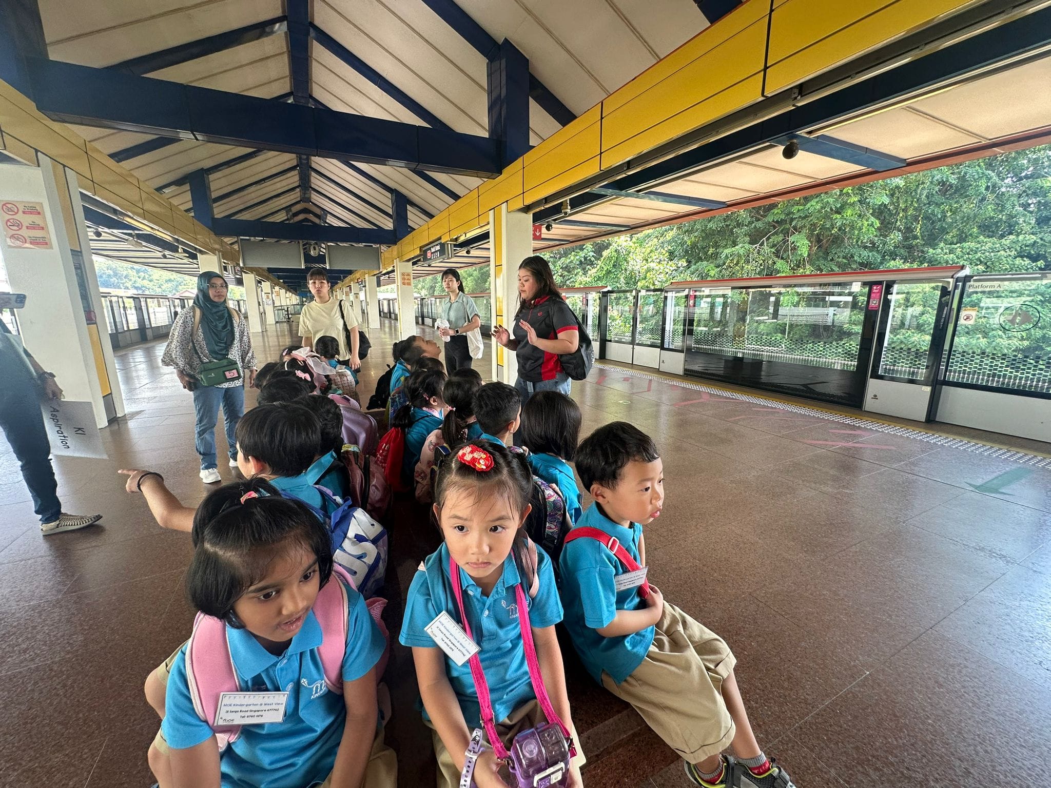 A group of young children in blue uniforms with backpacks lines up on a train station platform with adult chaperones.