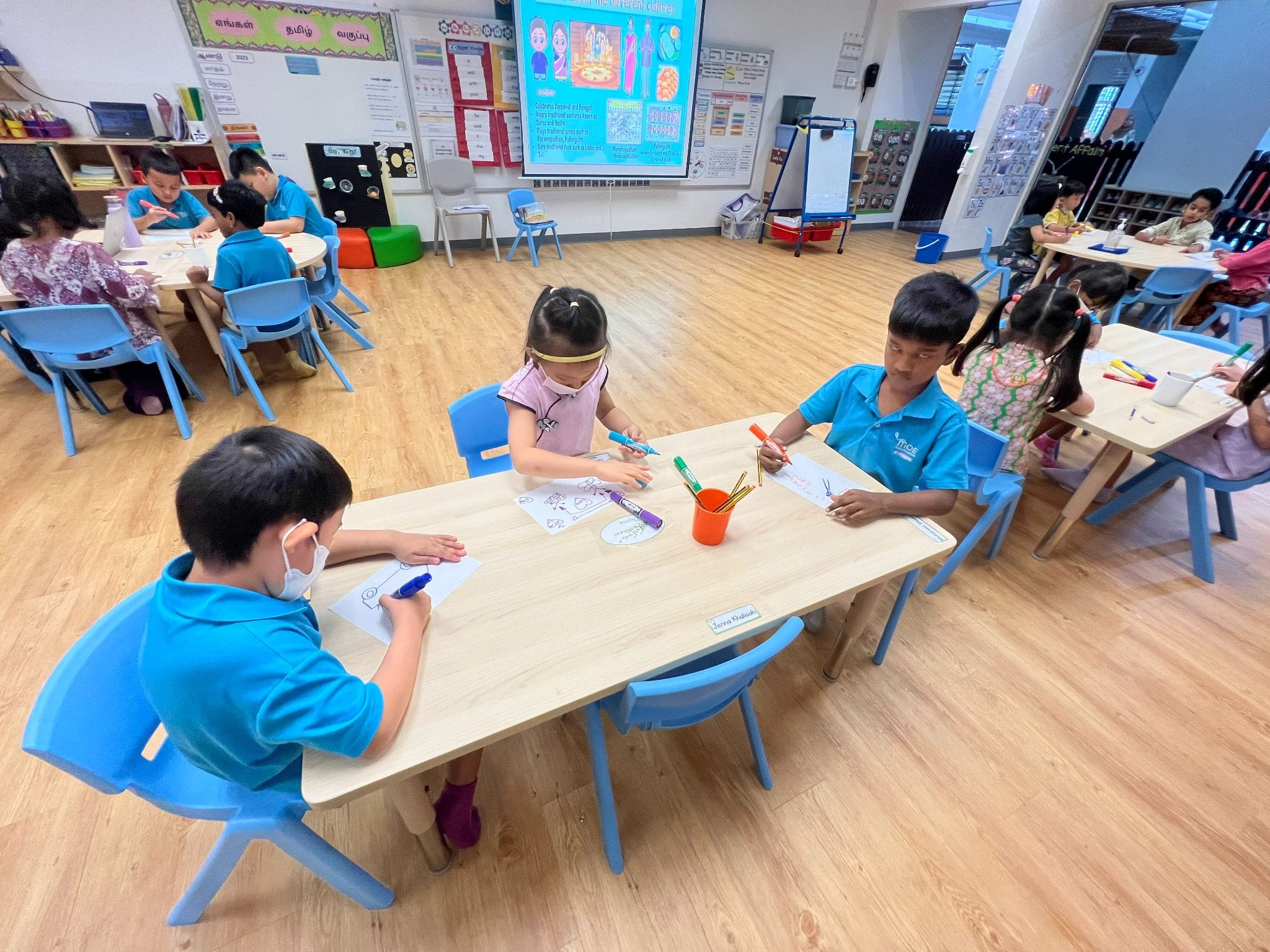 Young children in blue uniforms sit at tables in a bright classroom, drawing and coloring with markers on paper.