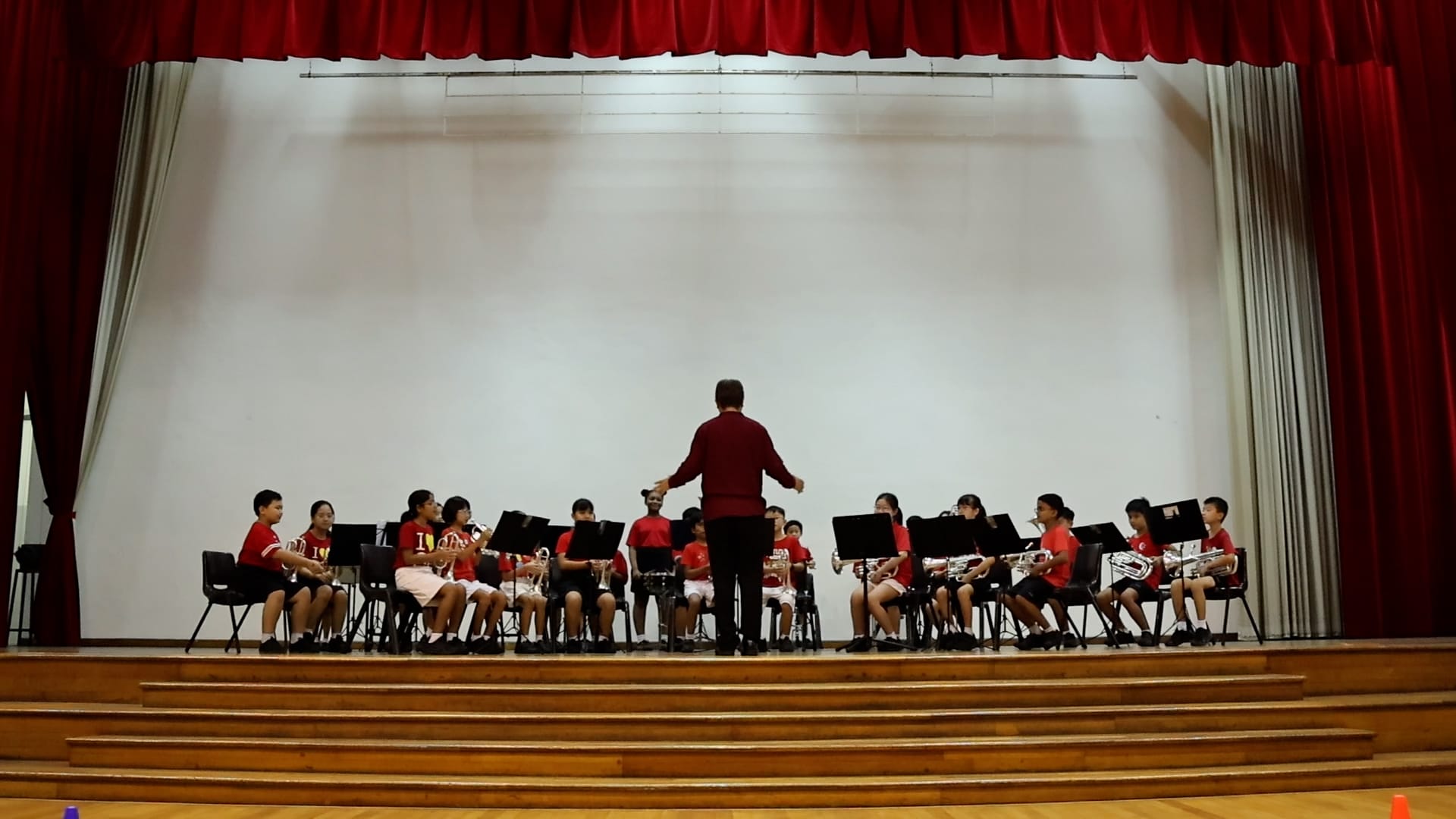 Children in red shirts play brass instruments on a stage, led by a conductor facing away from the camera.