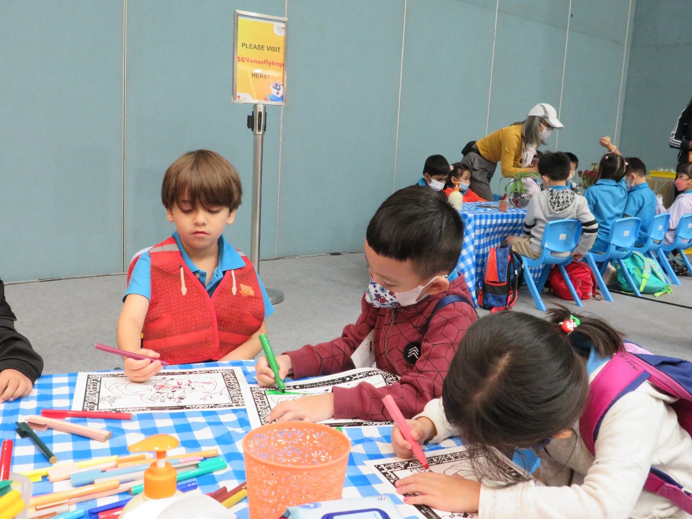 Children sit at a blue checkered table coloring printed sheets with markers at an indoor event.