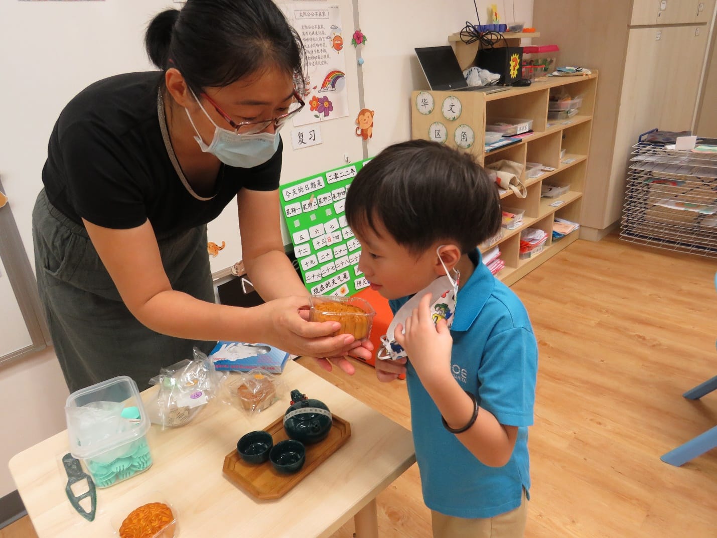 A masked teacher shows a mooncake to a young boy in a Chinese classroom with bookshelves and educational posters.