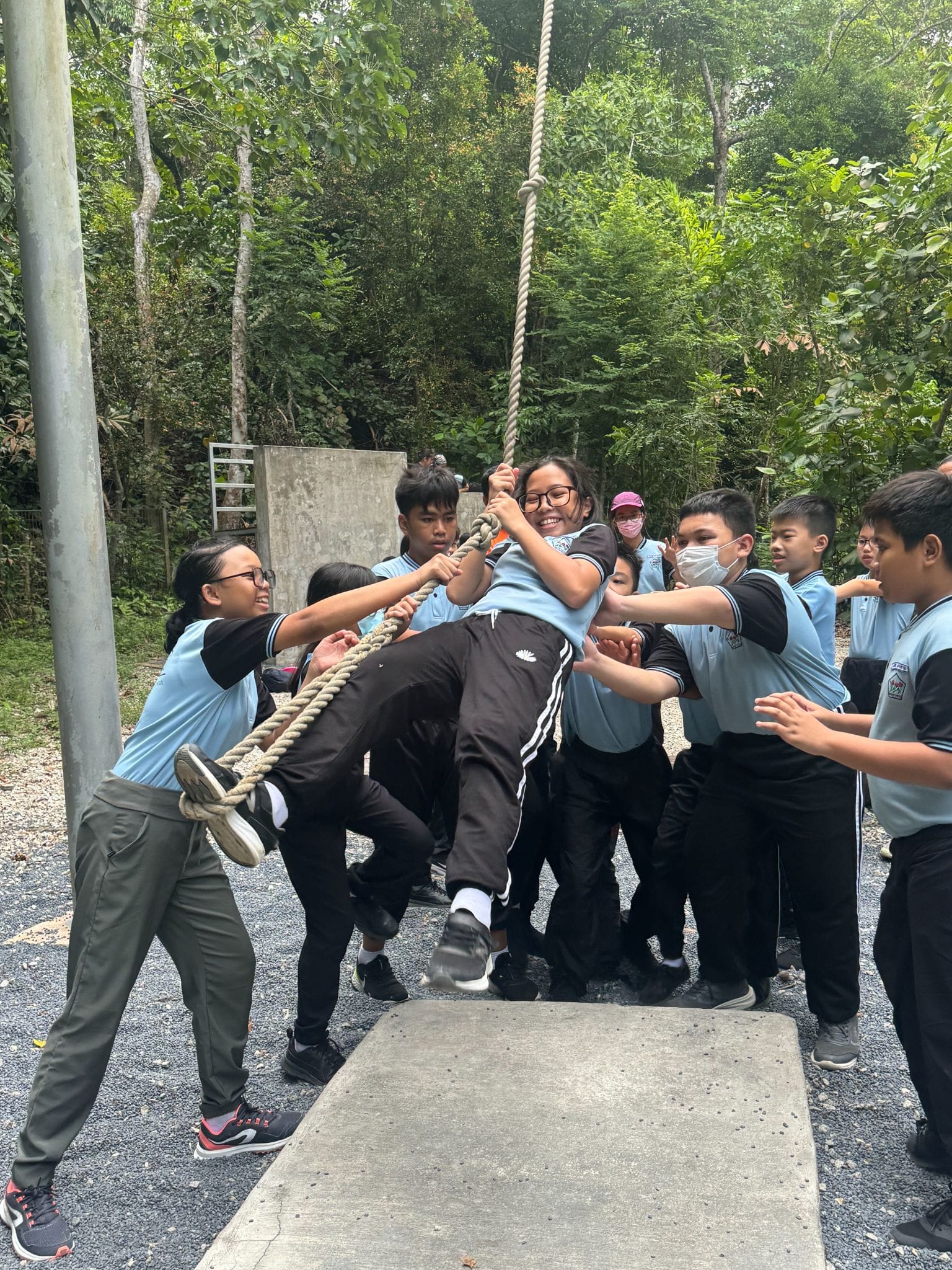 Students in light blue uniforms help a smiling girl climb a hanging rope outdoors near trees.