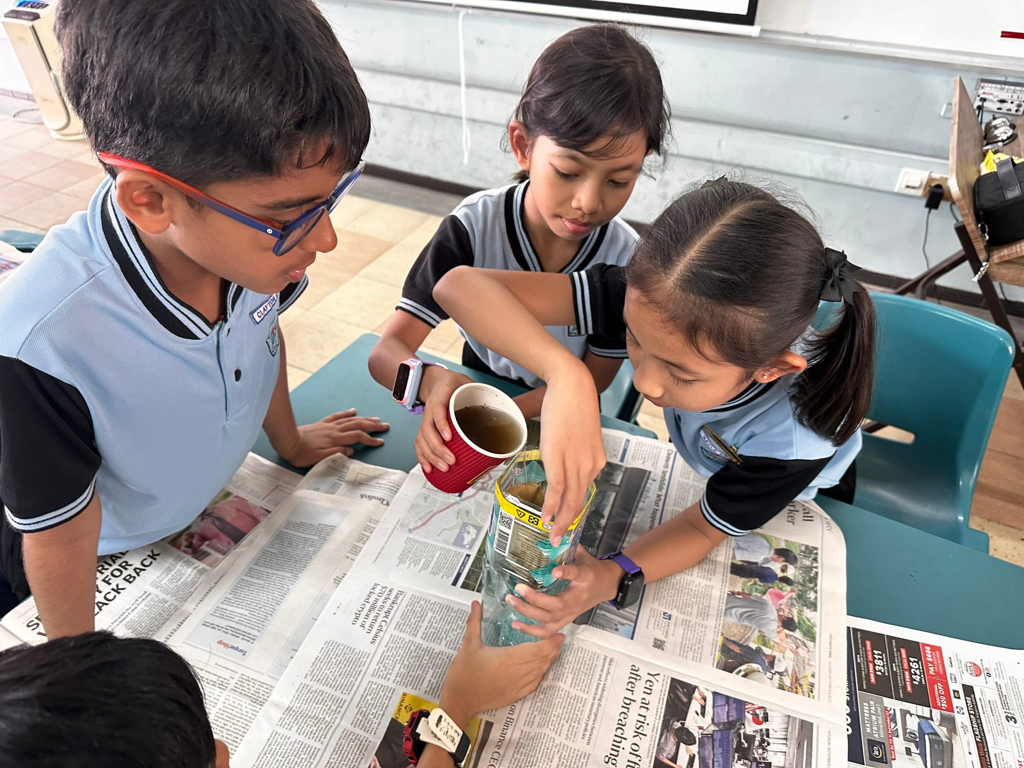 Three school children in uniforms pour liquid from a cup into a bottle on a newspaper-covered table.