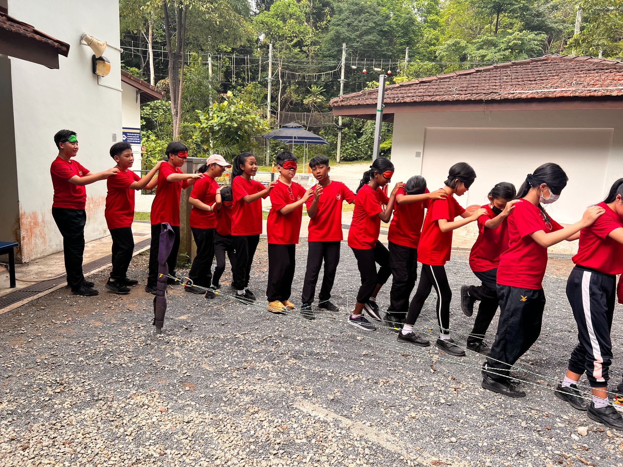 Children in red shirts form a chain, some blindfolded, stepping over a wire obstacle on a gravel outdoor area.