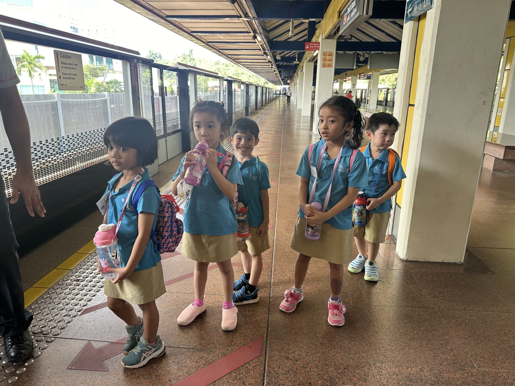 Five young children in matching blue uniforms with backpacks and water bottles stand on a train station platform.