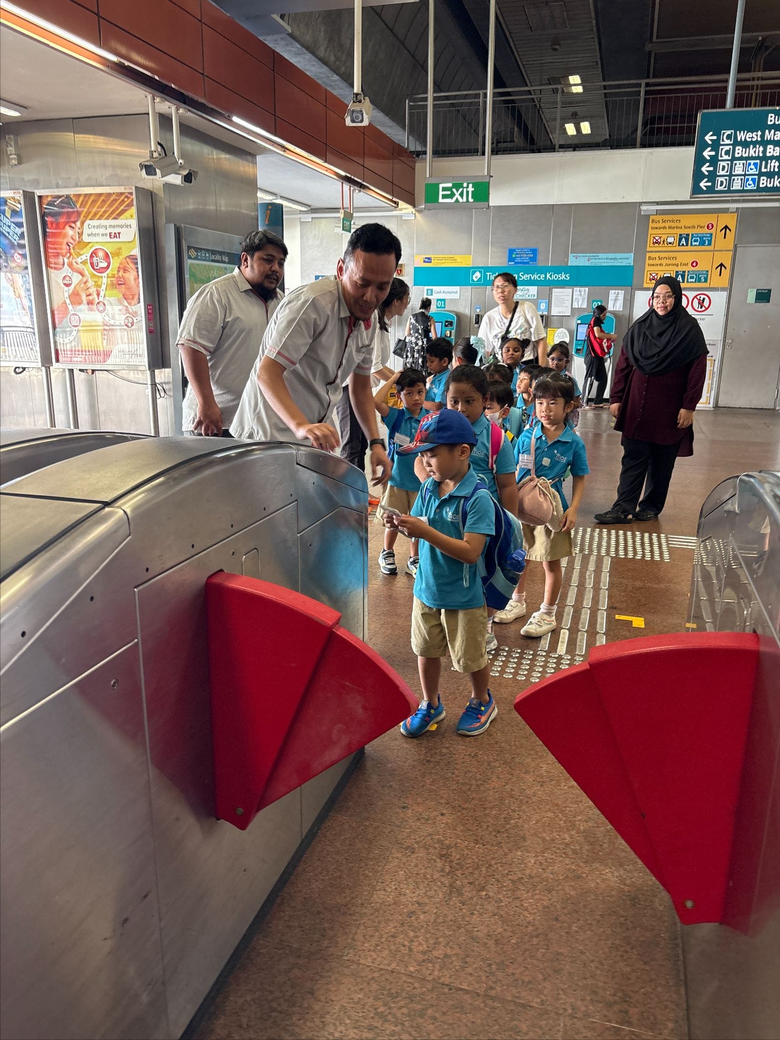 Young children in blue uniforms pass through red fare gates at a Singapore MRT station, guided by adults.