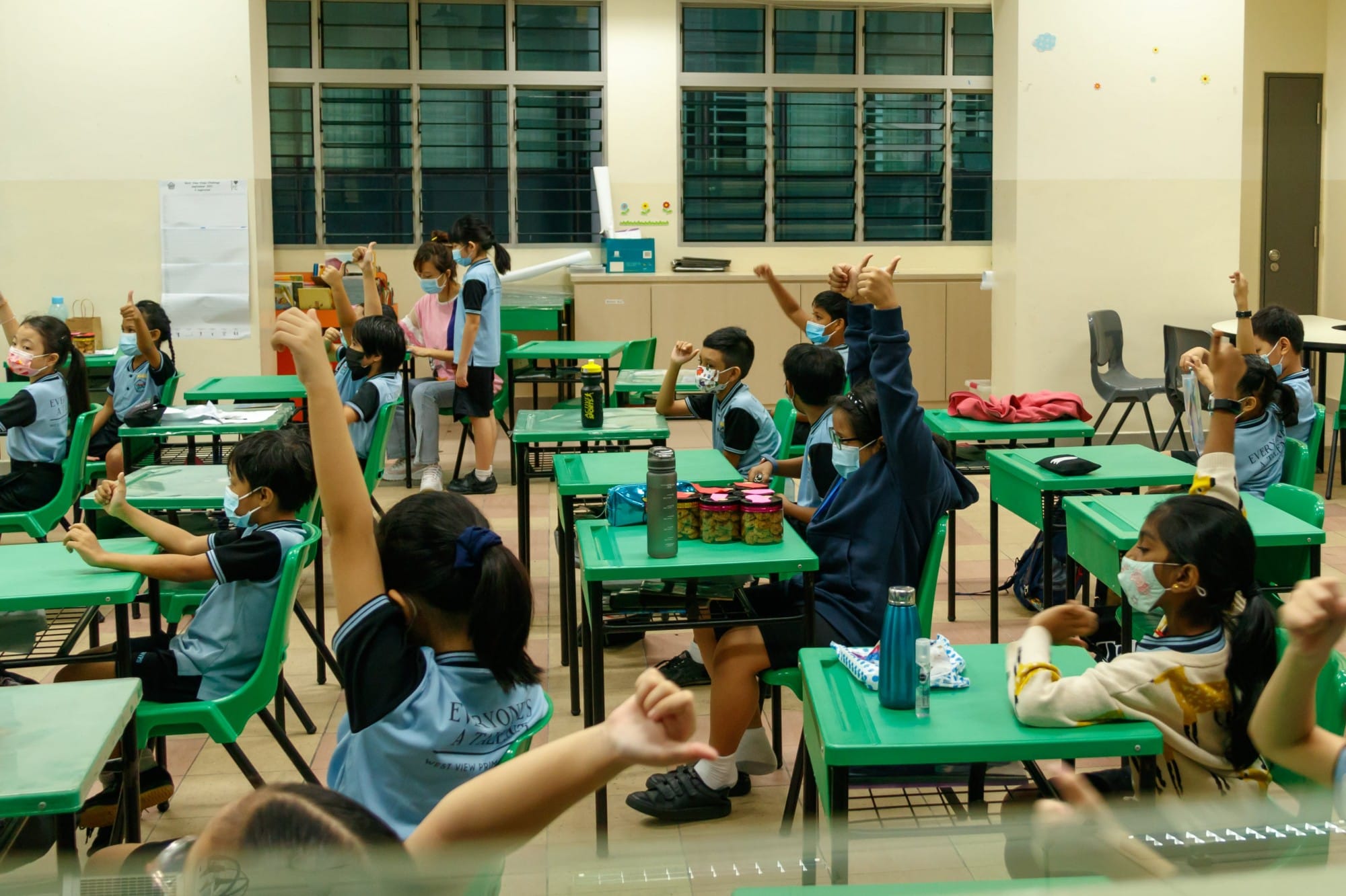 Young students in blue uniforms and face masks sit at green desks, raising hands and giving thumbs up in a classroom.
