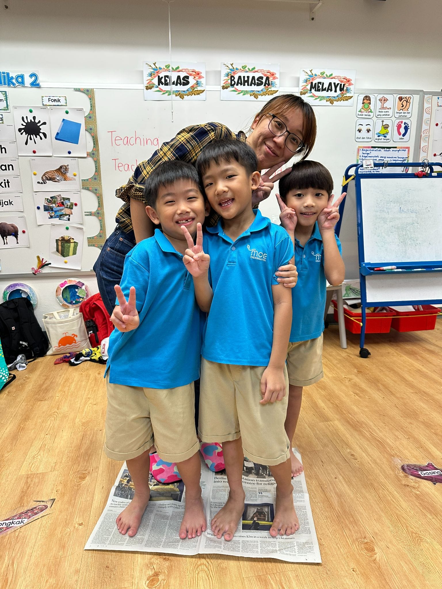 Three boys in blue uniforms make peace signs with a smiling teacher in a Malay language classroom.