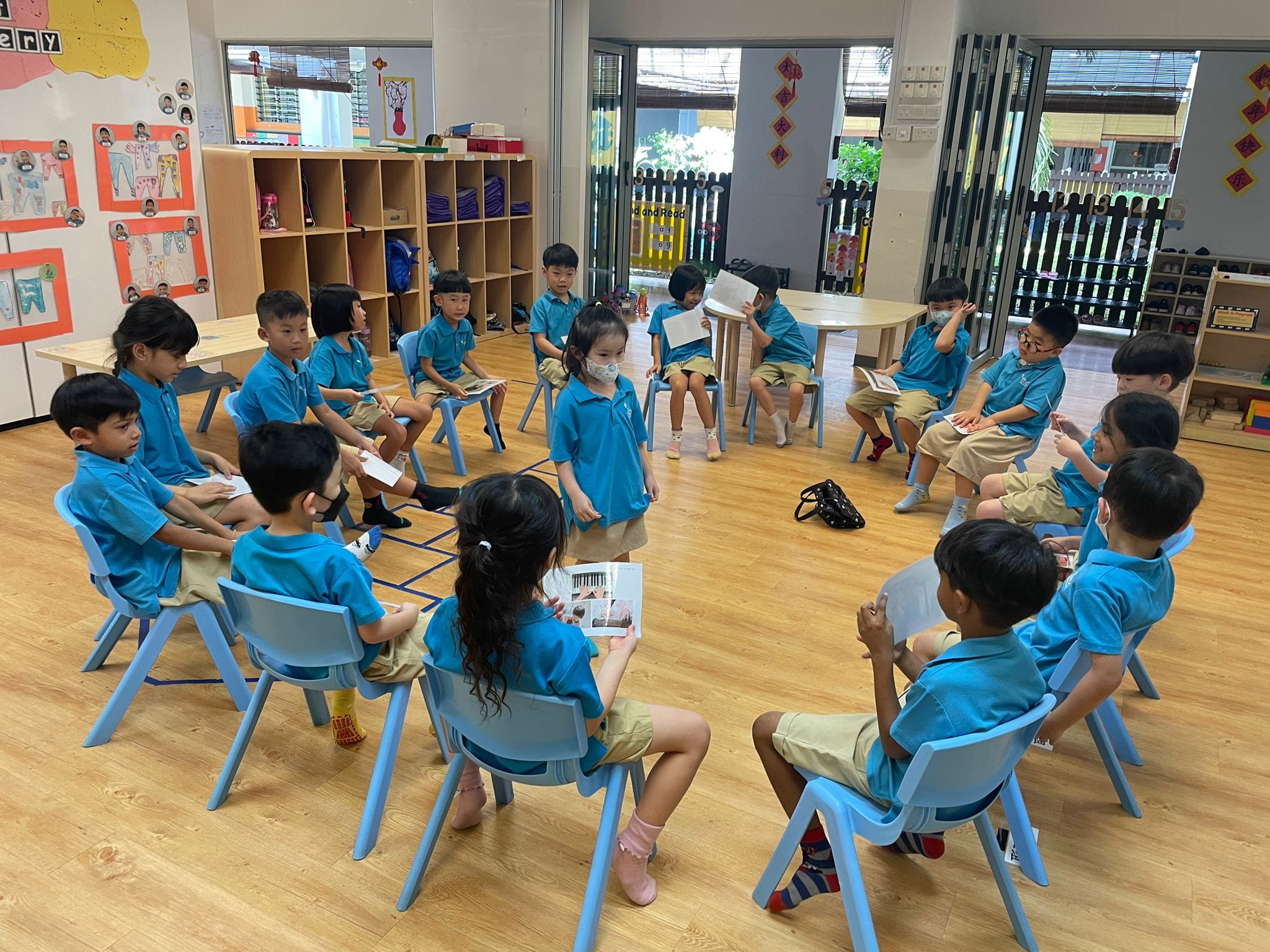 Young children in blue uniforms sit in a circle on small chairs in a bright classroom, holding books during a reading activity.