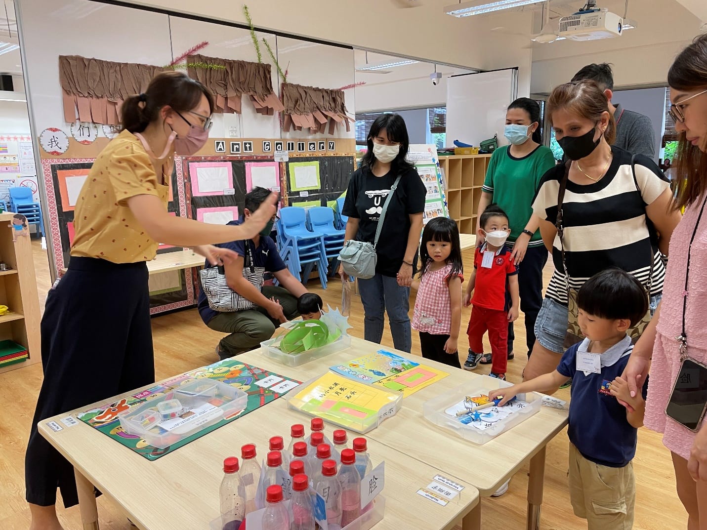 A teacher in a yellow top demonstrates activities to masked parents and young children around a table in a colorful classroom.