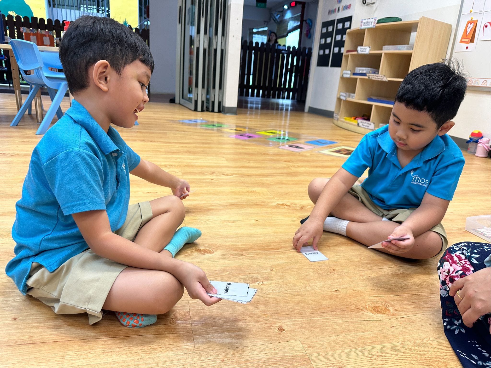 Two young boys in blue uniforms sit cross-legged on a classroom floor, playing with word flashcards.