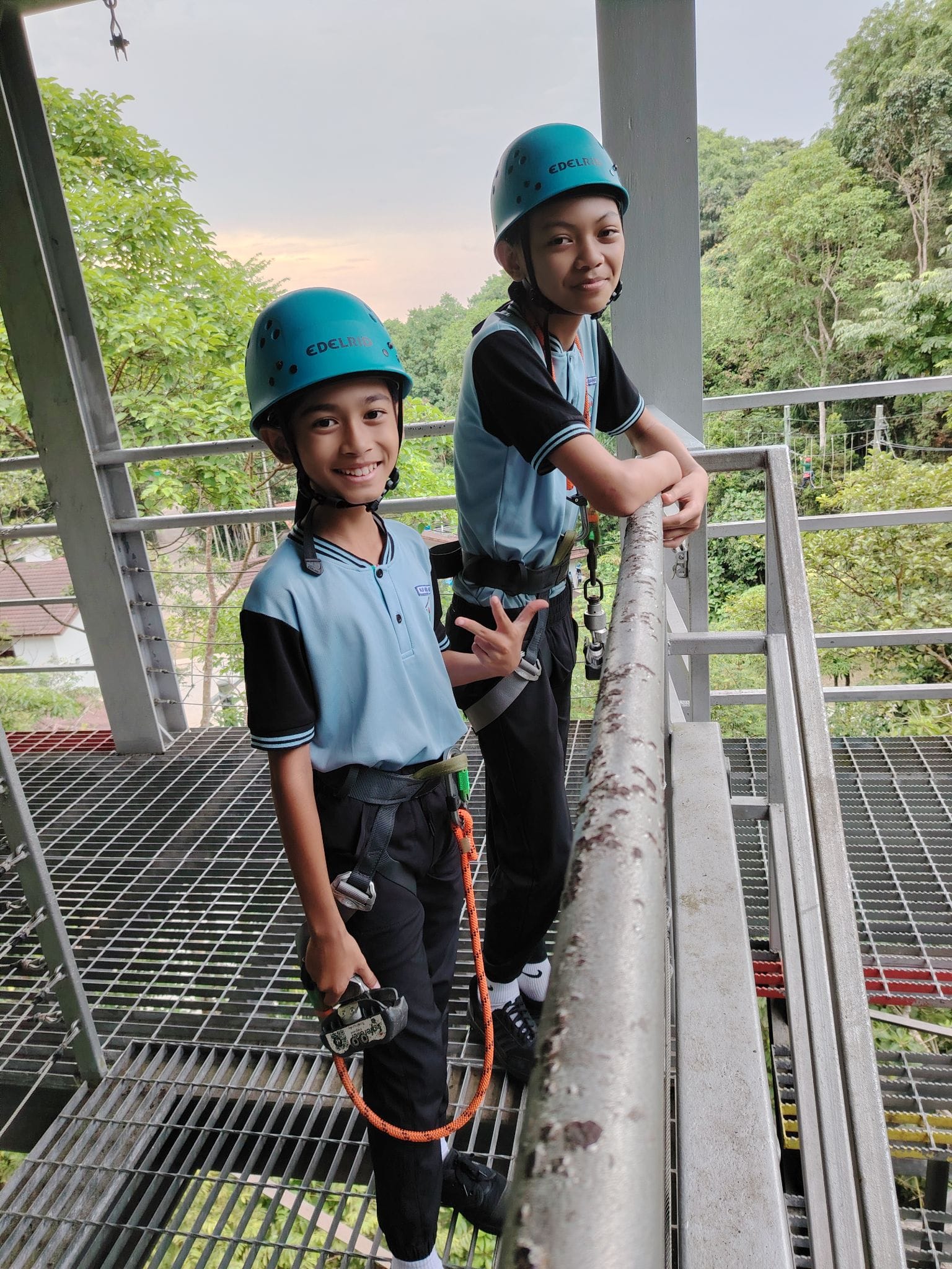 Two children in teal helmets and harnesses stand smiling on a metal grated platform surrounded by green trees.