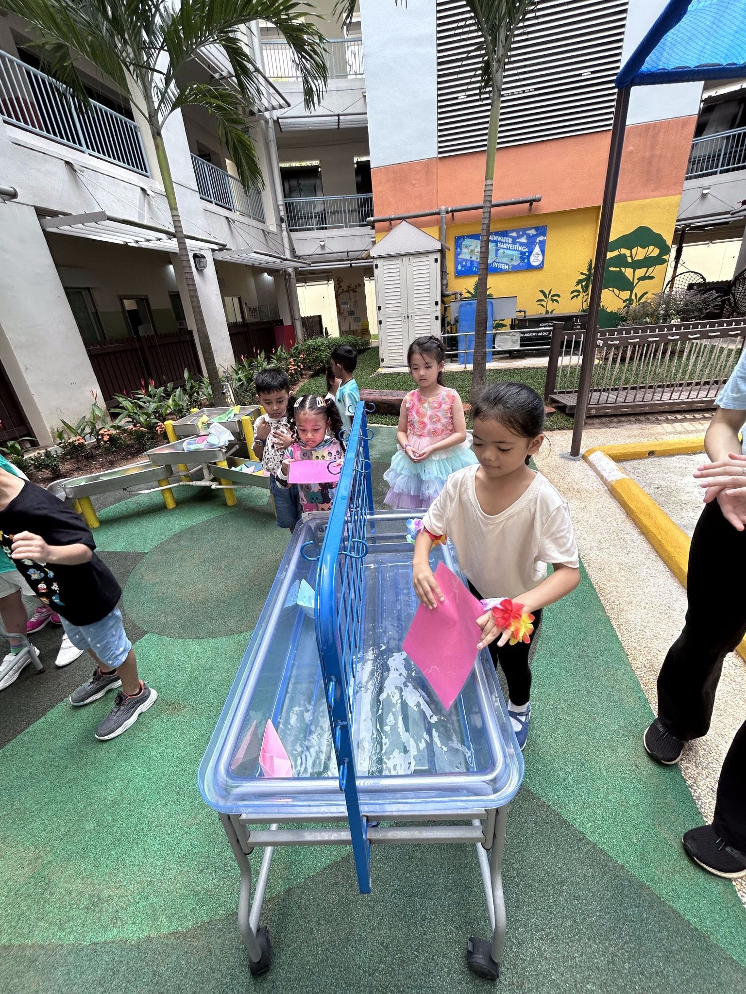 Several young children play around a clear water table with pink paper boats in an outdoor school courtyard.