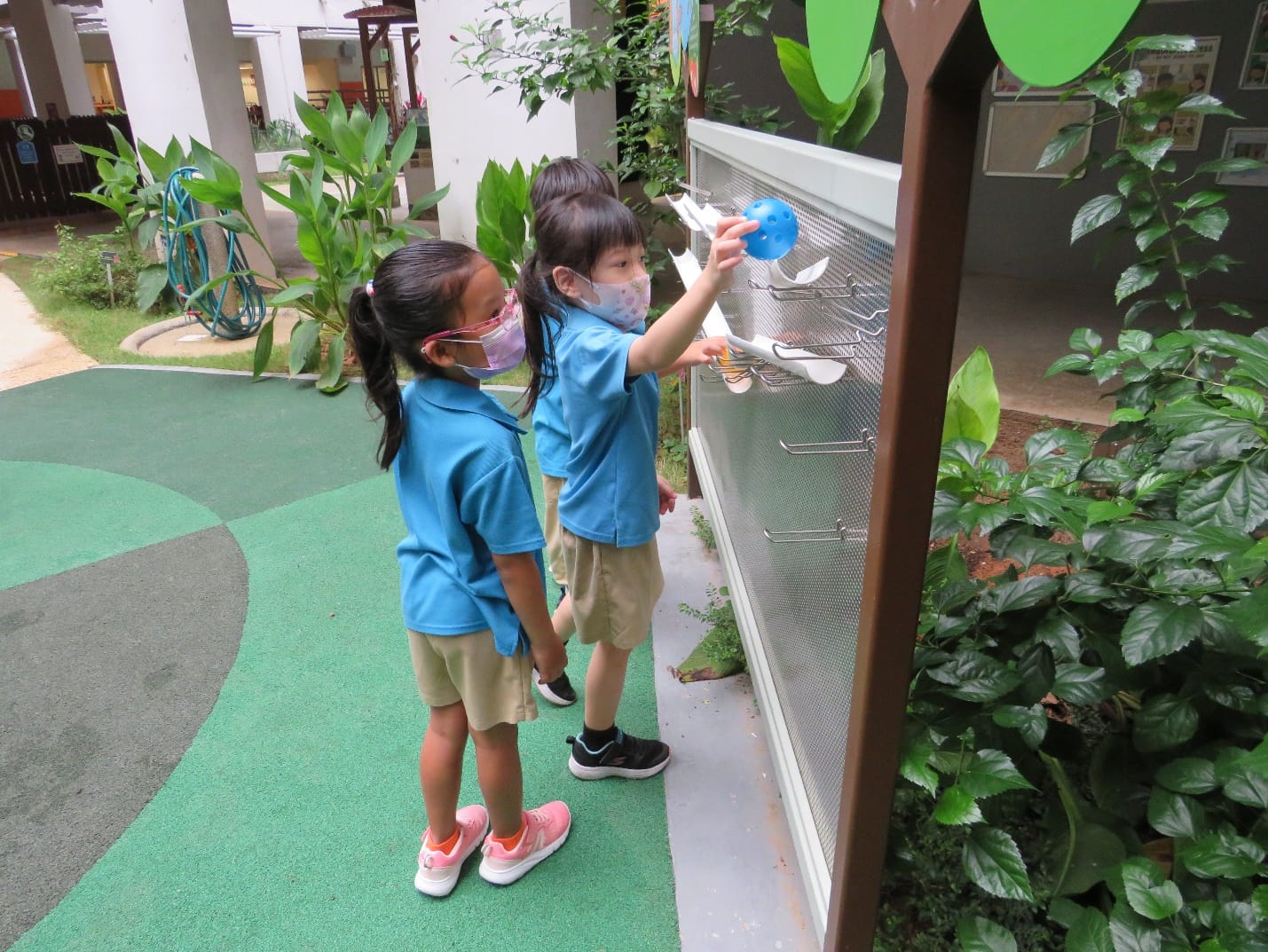 Two young children in blue school uniforms and face masks play with an outdoor wall panel holding a blue ball.