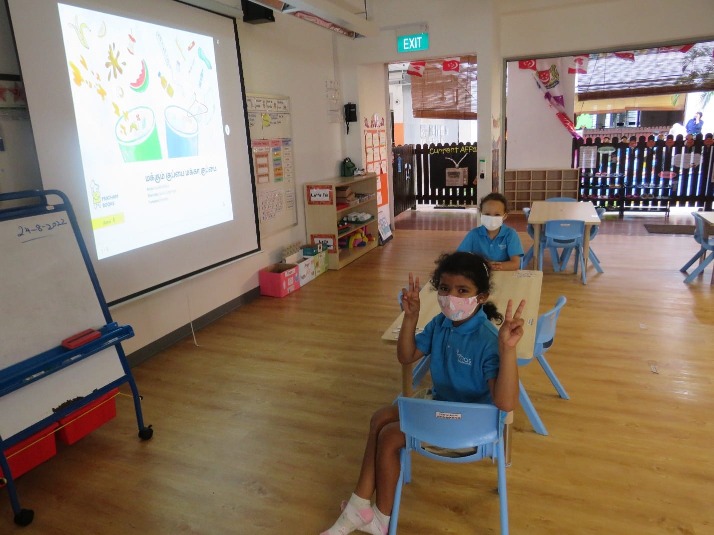 Two masked children in blue uniforms sit in a classroom facing a projected screen with Tamil text.