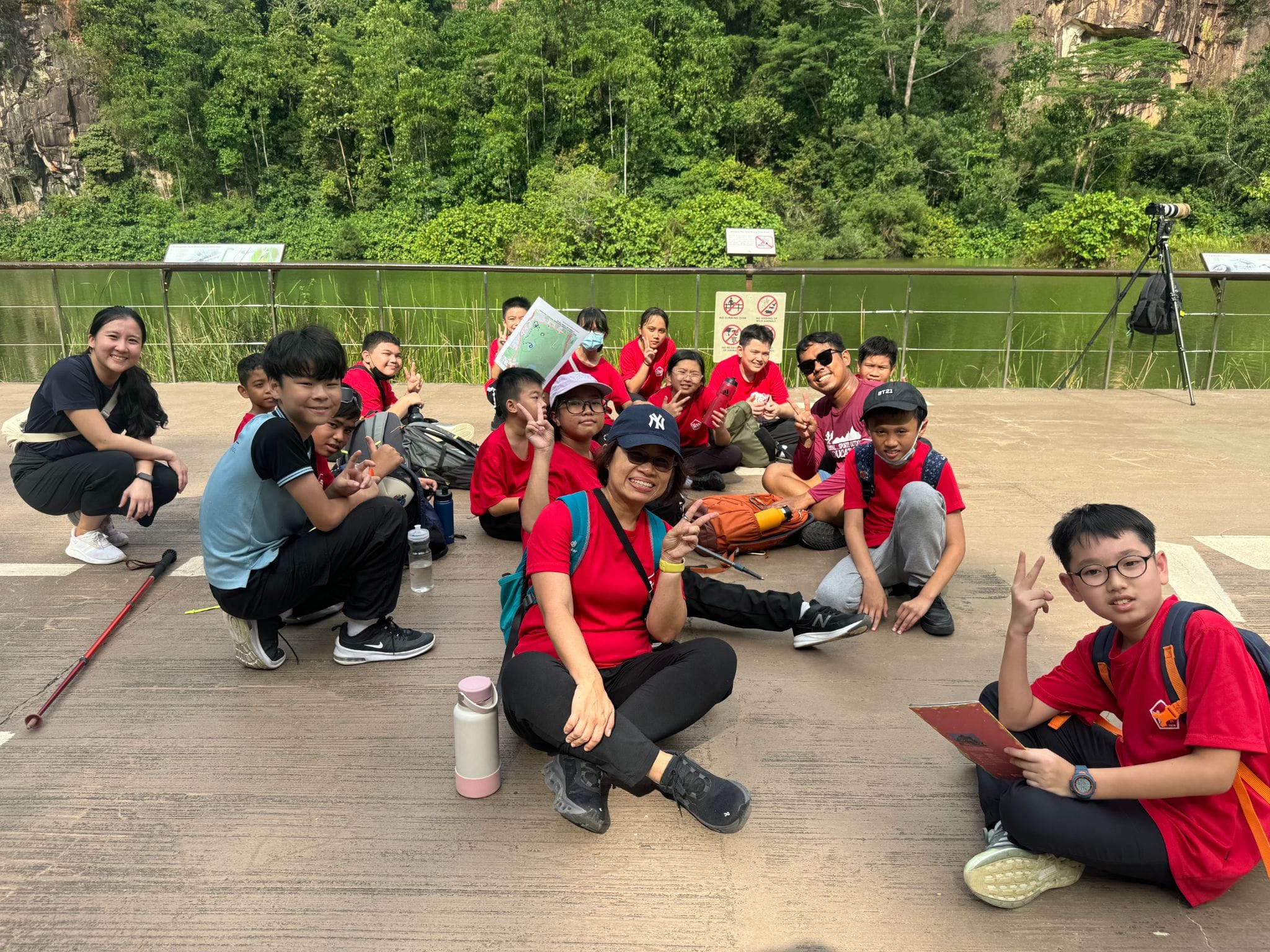 A group of children and adults in red shirts pose with peace signs near a lake surrounded by rocky cliffs and trees.
