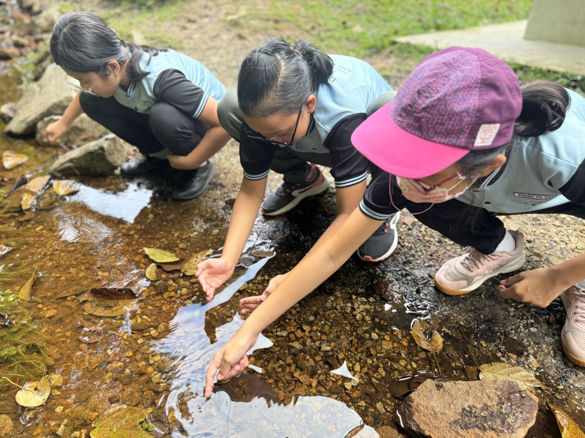 Three children in blue uniforms crouch beside a shallow rocky stream, reaching their hands into the water.