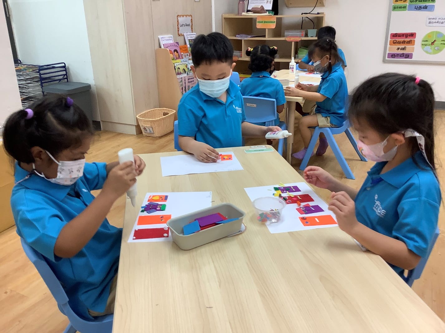 Three masked children in blue uniforms do a colorful craft activity at a table in a classroom.