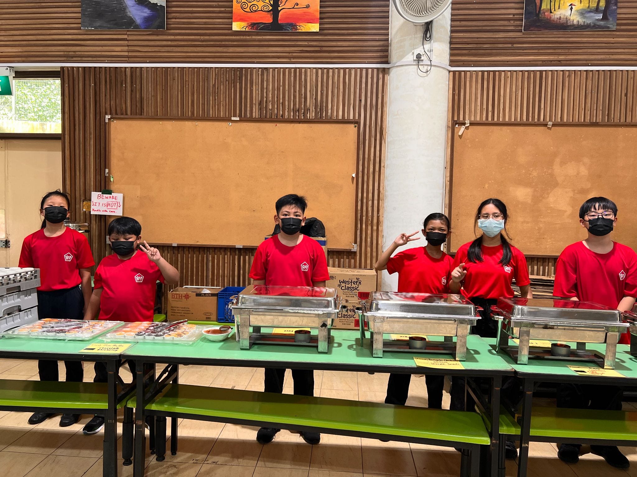 Six students in red shirts and black masks stand behind a buffet table with chafing dishes and food trays.