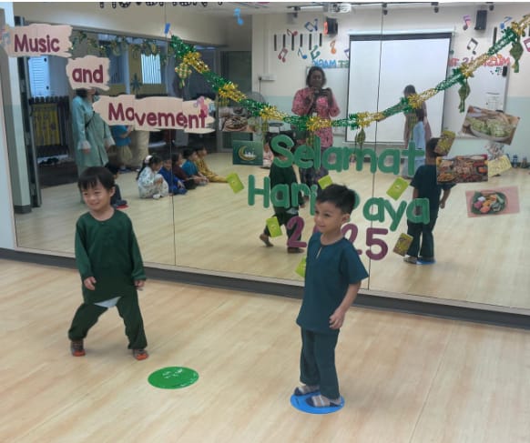 Two young children in green outfits stand in a dance studio with a Selamat Hari Raya 2025 decorated mirror.