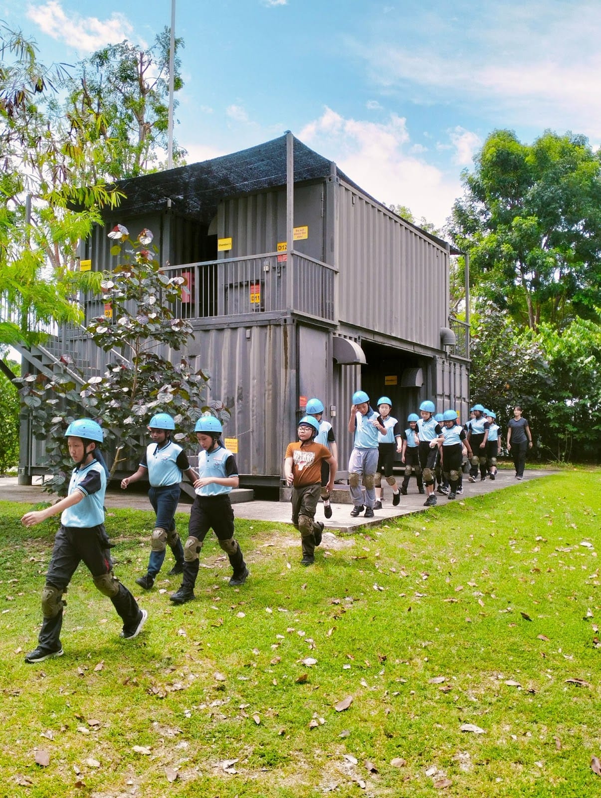 A group of children wearing blue helmets and knee pads walk on grass past a two-story shipping container structure.