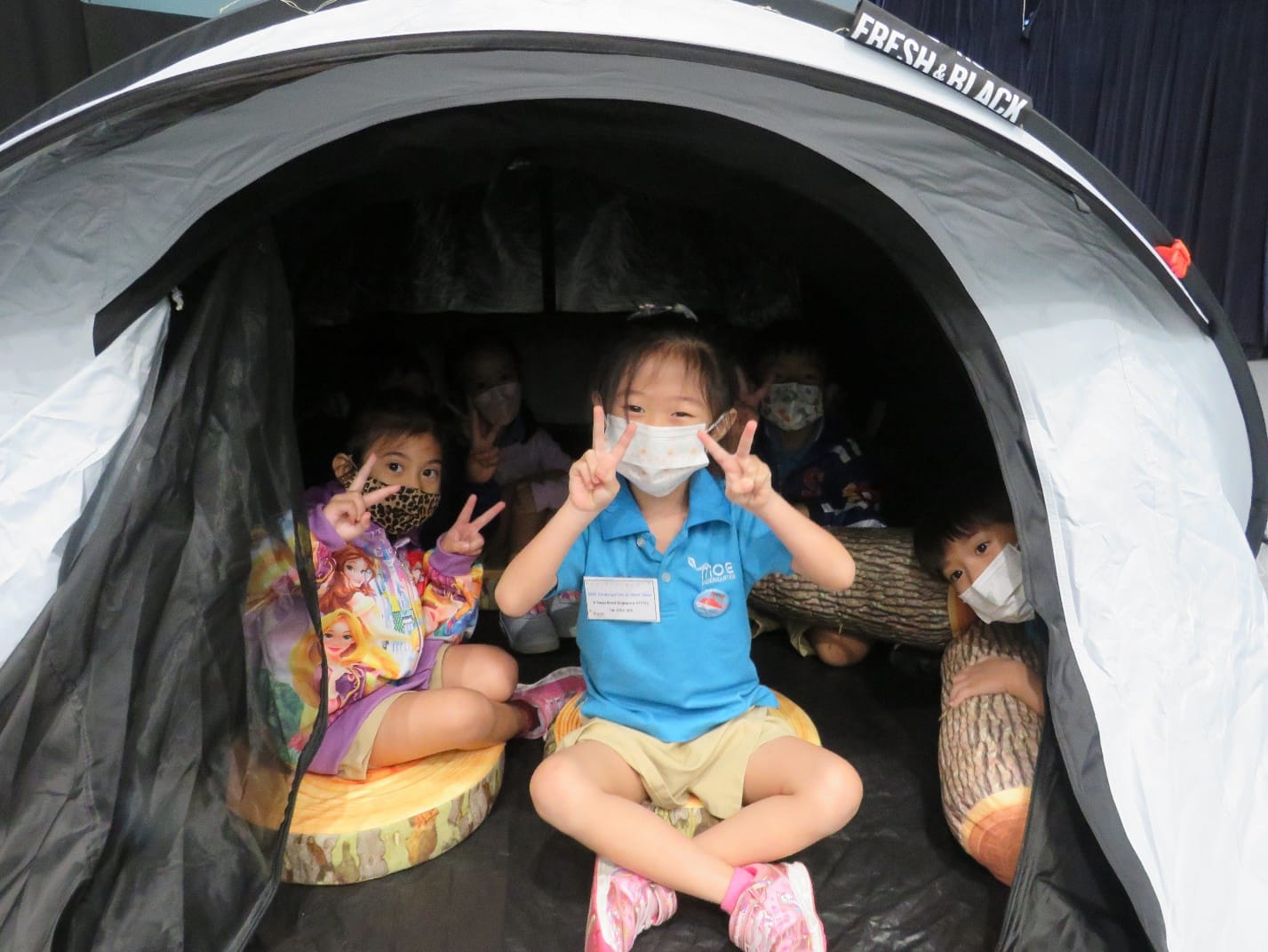 Several young children wearing face masks sit inside a small dome tent, smiling and making peace signs.