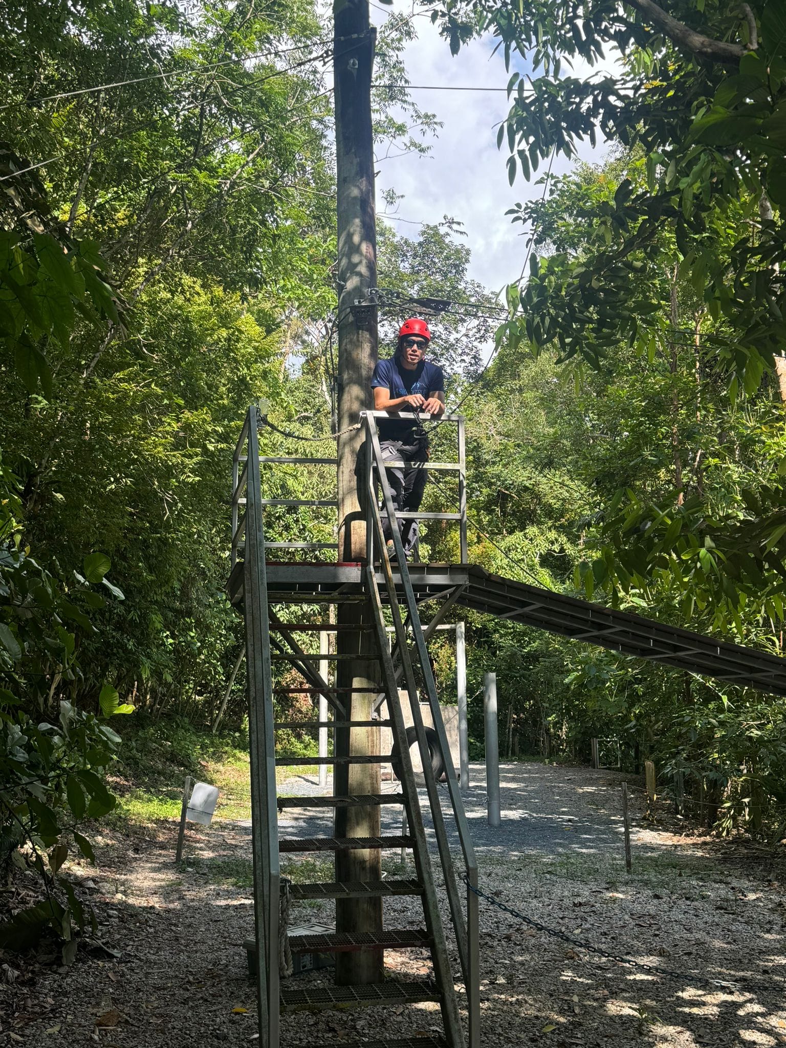 Person wearing a red helmet stands on a tall metal zipline platform surrounded by dense green trees.